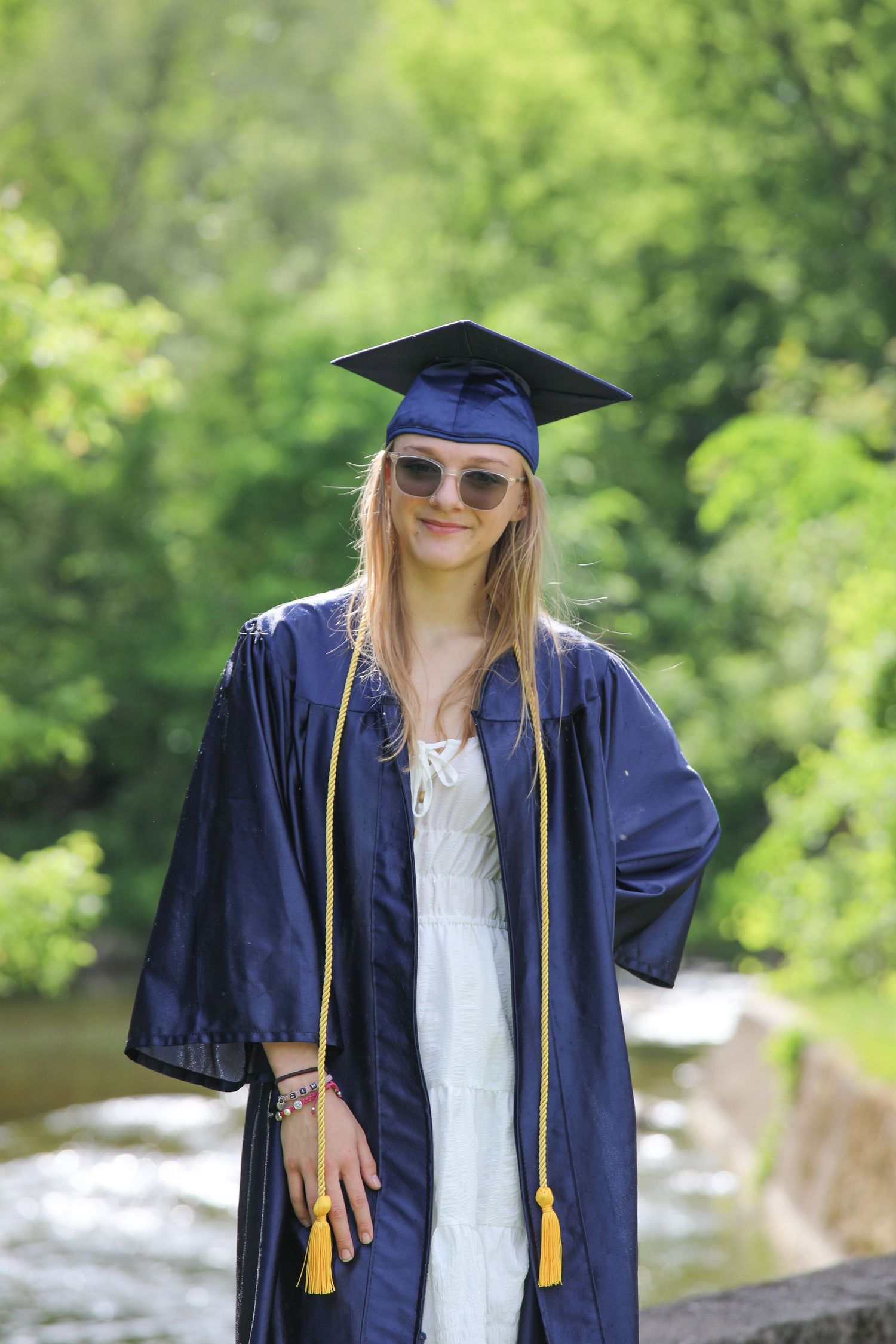 Young woman in navy blue graduation cap and gown with yellow tassel, smiling outdoors near a creek.