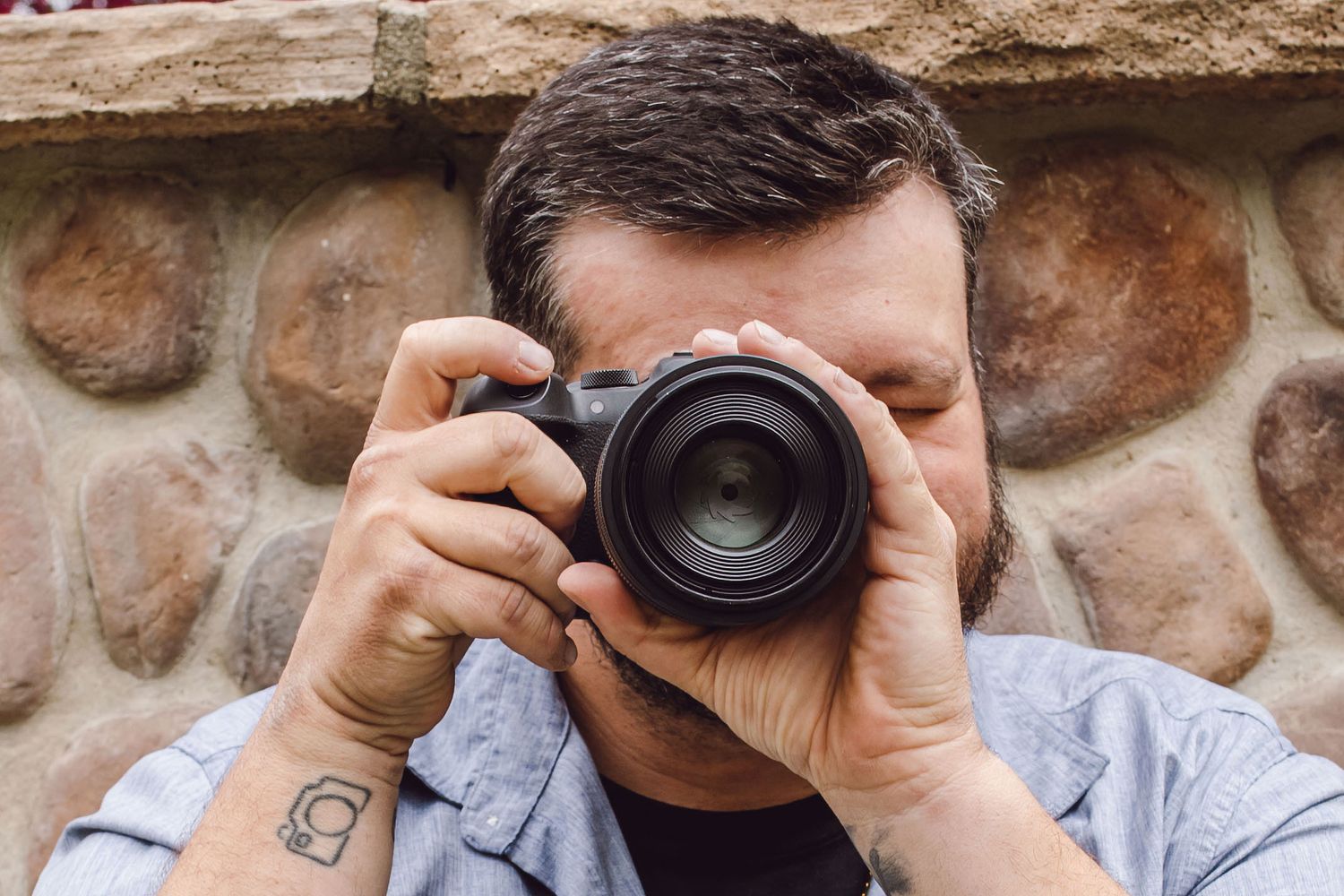 A bearded man holds a DSLR camera up to his eye, photographing against a rustic stone wall background.