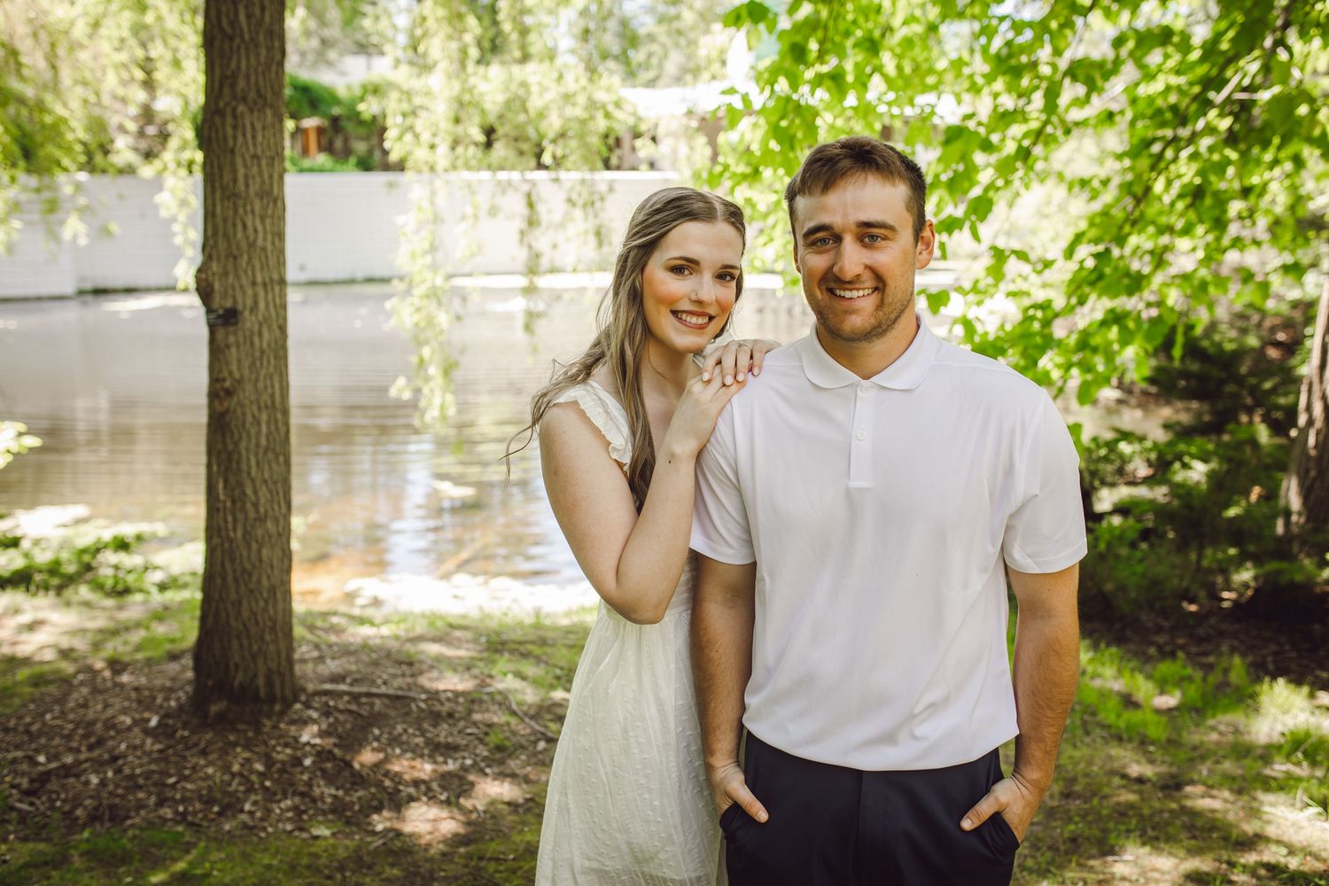 Smiling couple posing outdoors near a pond surrounded by lush green trees on a sunny day.