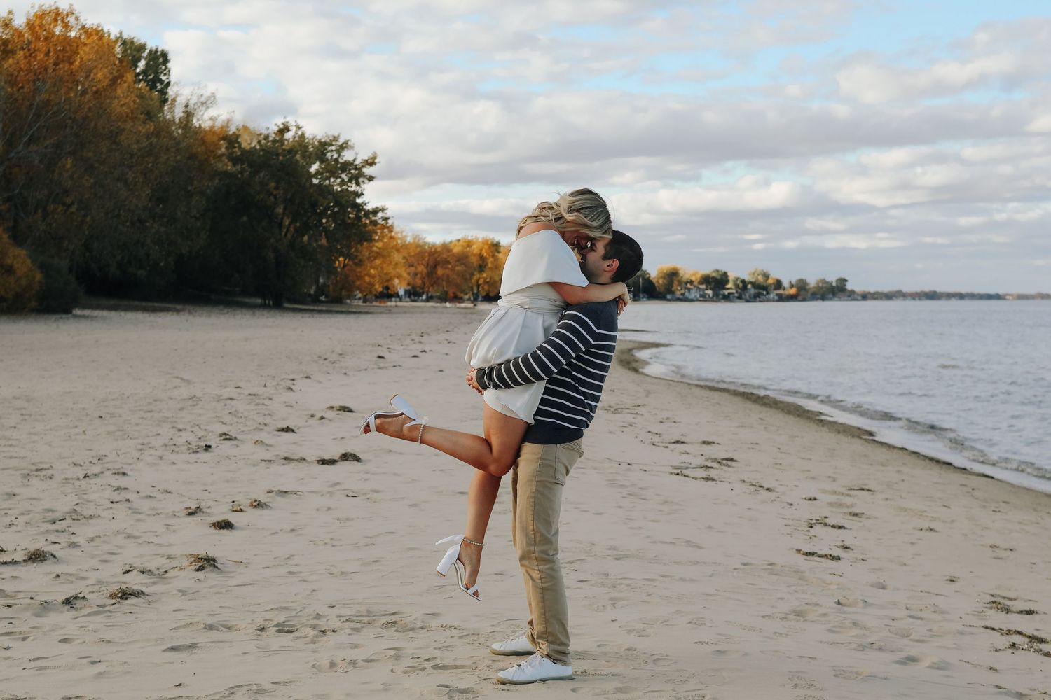 A playful lift pose on a beach with autumn trees and water in the background.