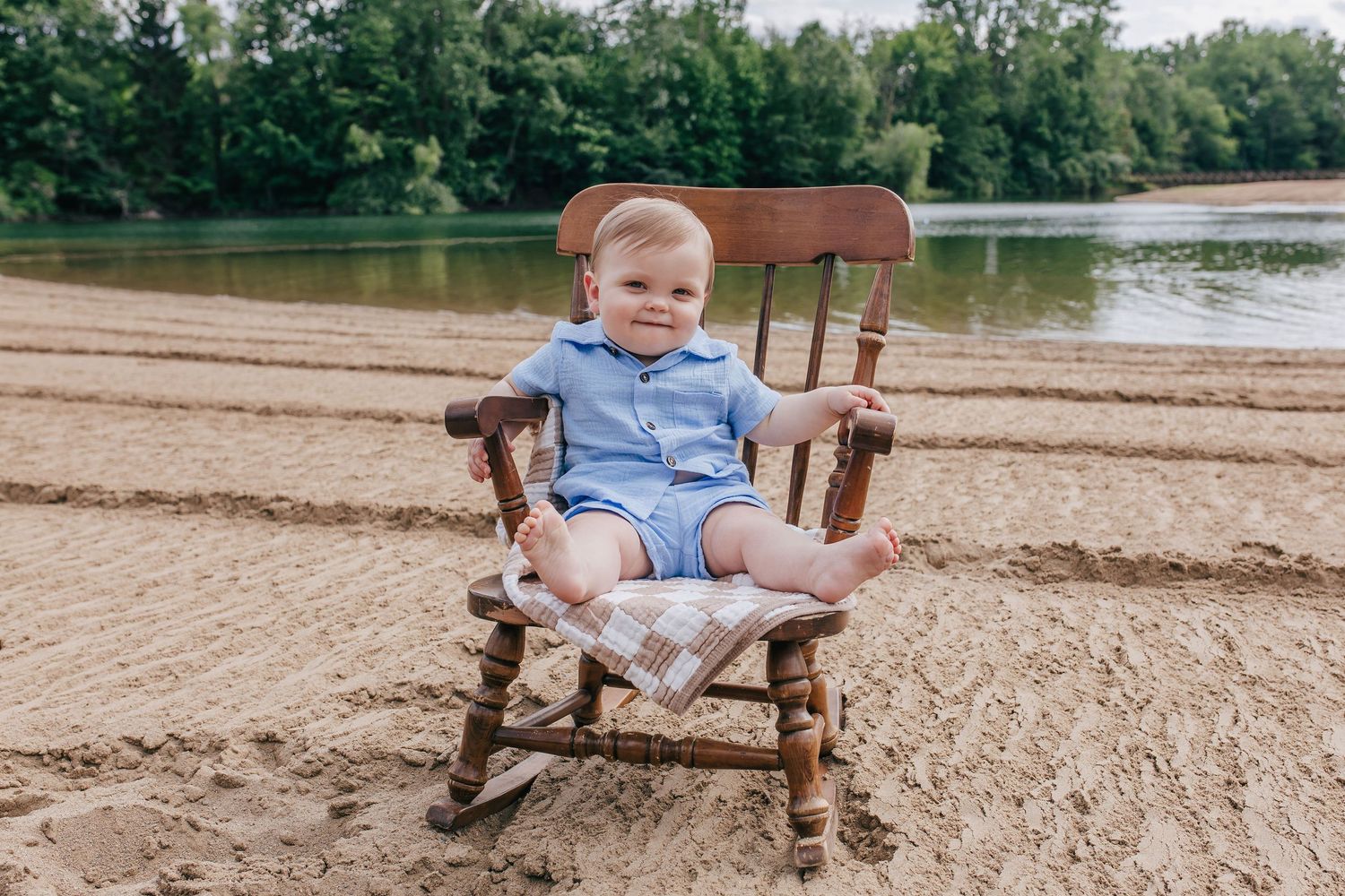 A smiling baby in a blue romper sits in a wooden rocking chair on a sandy beach by a river.