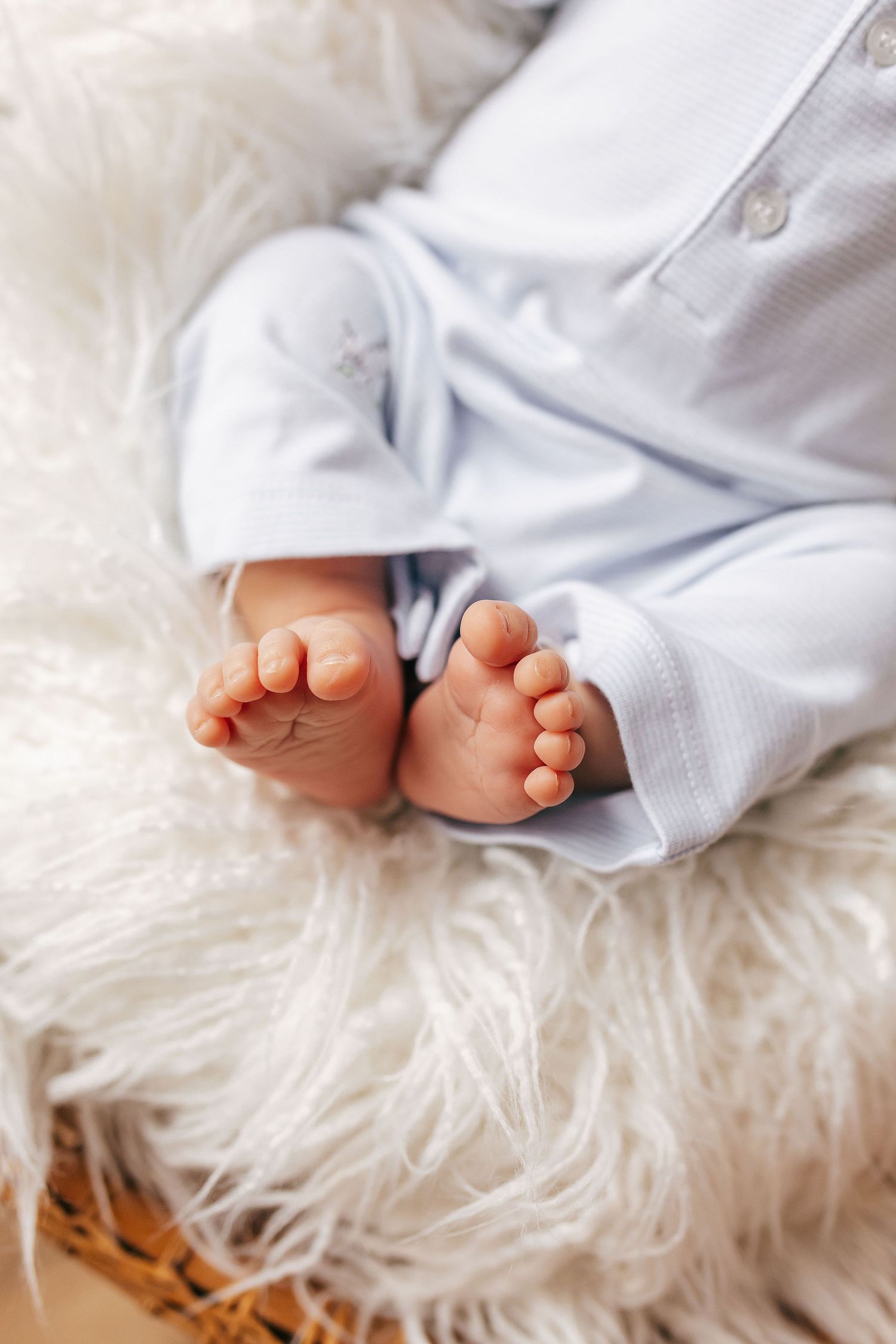 Close-up of tiny newborn baby feet in light blue onesie resting on soft white fluffy blanket.