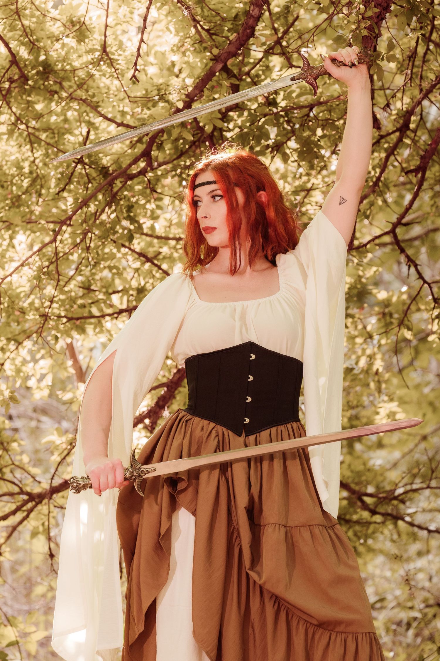 Red-haired woman in medieval costume holding two swords outdoors among green trees.