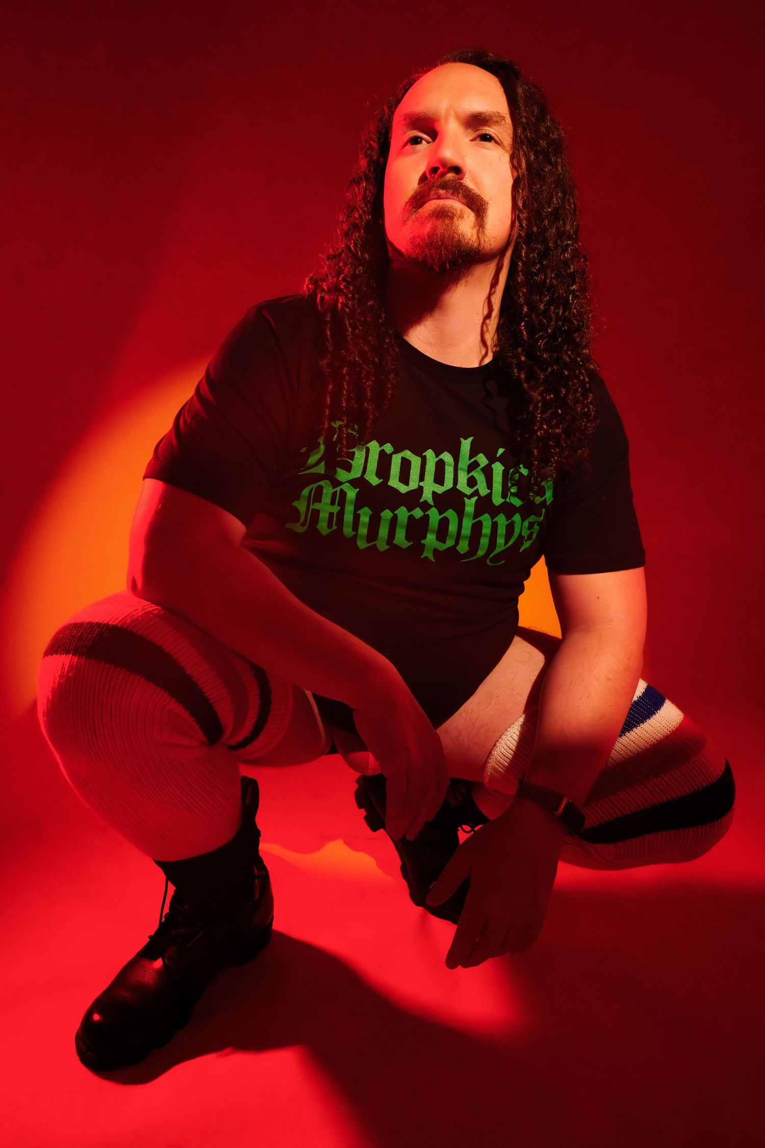 Man with curly hair wearing a Dropkick Murphys black t-shirt sitting against a red background.
