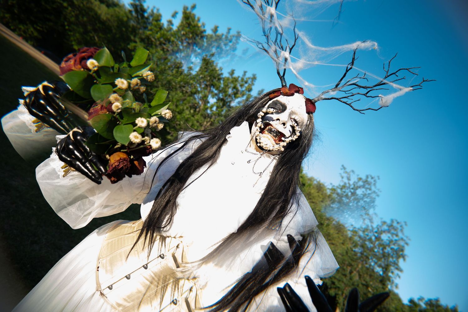 Person in elaborate white costume with skull face paint holding flowers and branches outdoors.
