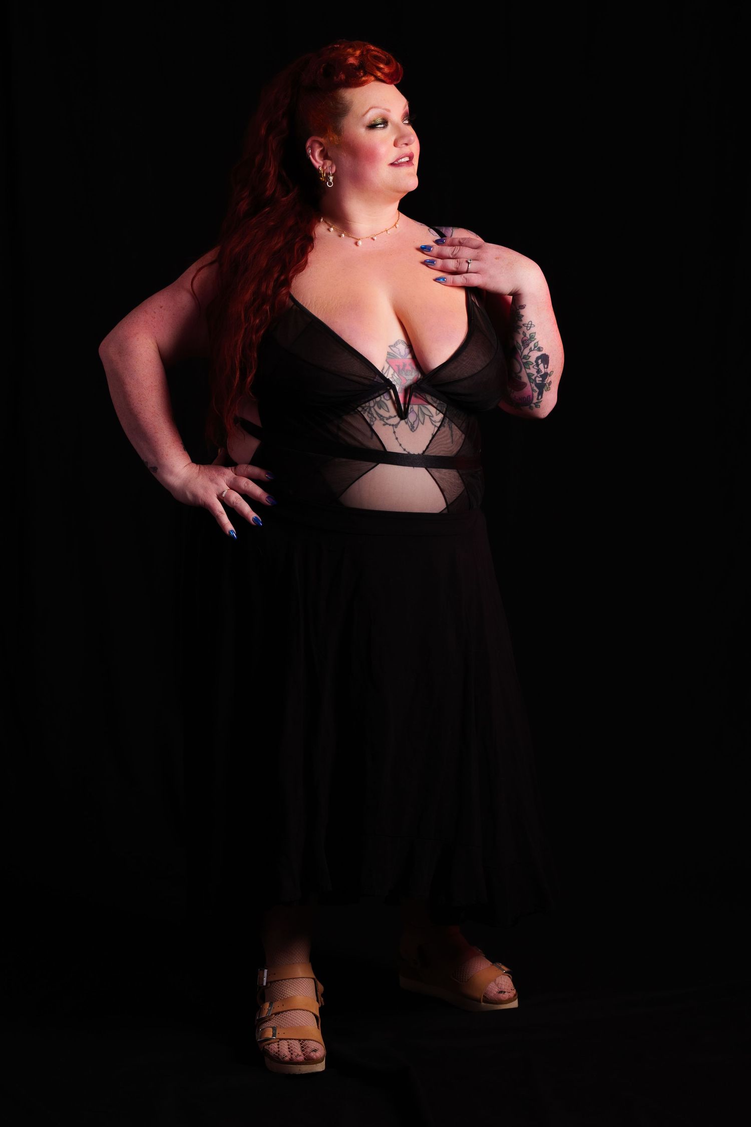Plus-size woman with red hair in black maxi dress with sheer panels, tattoos, posing confidently on dark background.