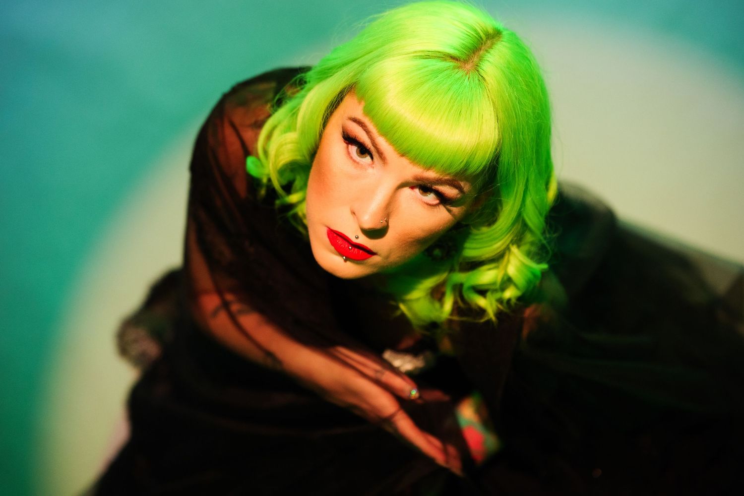 Woman with vibrant neon green wavy wig and red lips poses dramatically against a teal background.