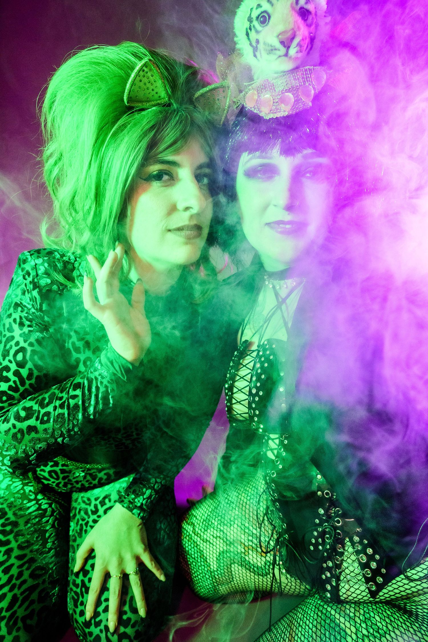 Two women in vibrant green and purple costumes pose together surrounded by colorful smoke.