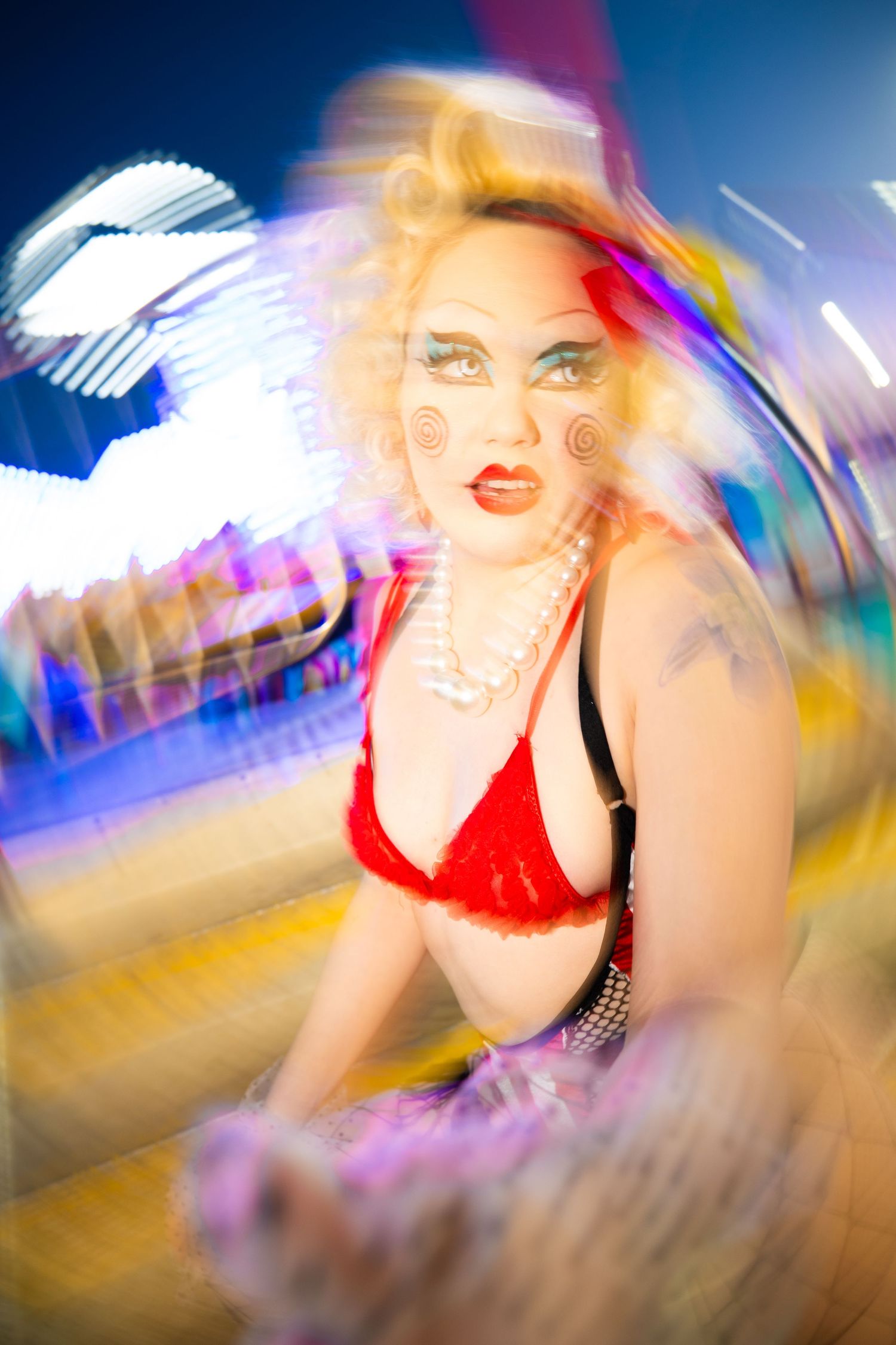Blonde woman in red bra with clown makeup at a colorful carnival with motion blur.