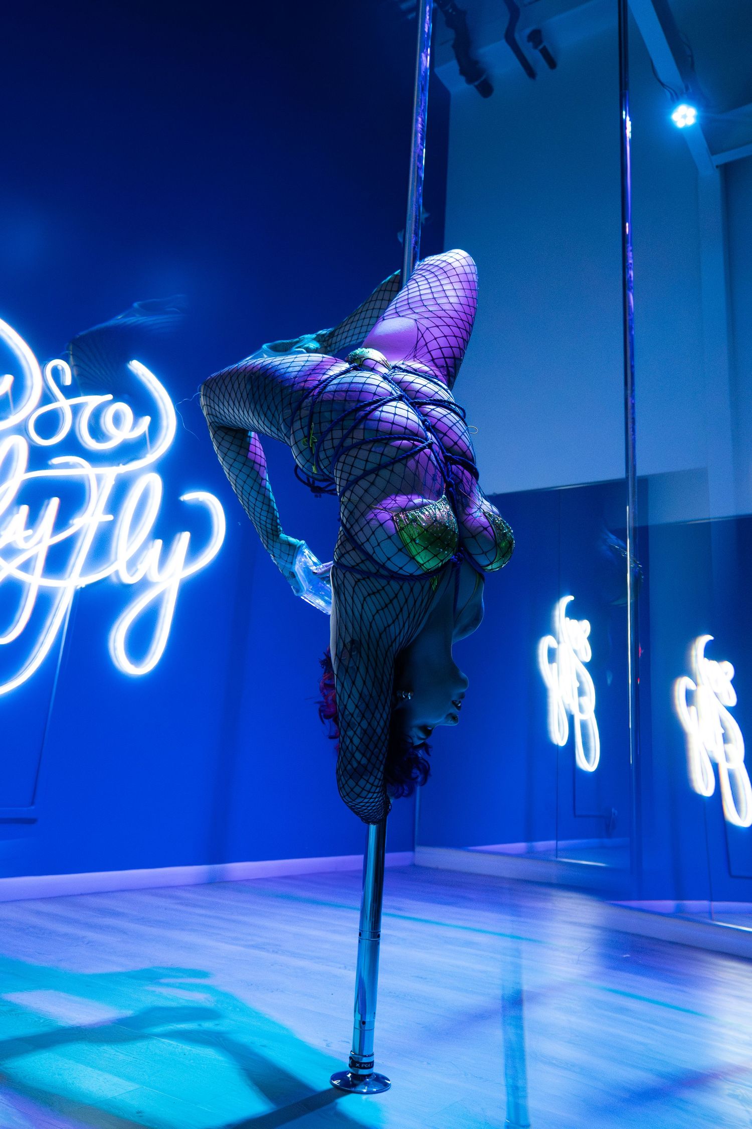 Pole dancer performing inverted trick under blue neon lights.