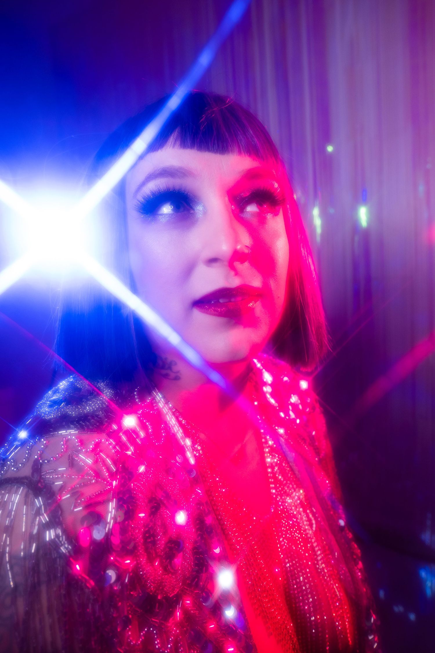 Woman in sequined jacket under neon pink and blue lights with lens flare in a vibrant nightclub setting.