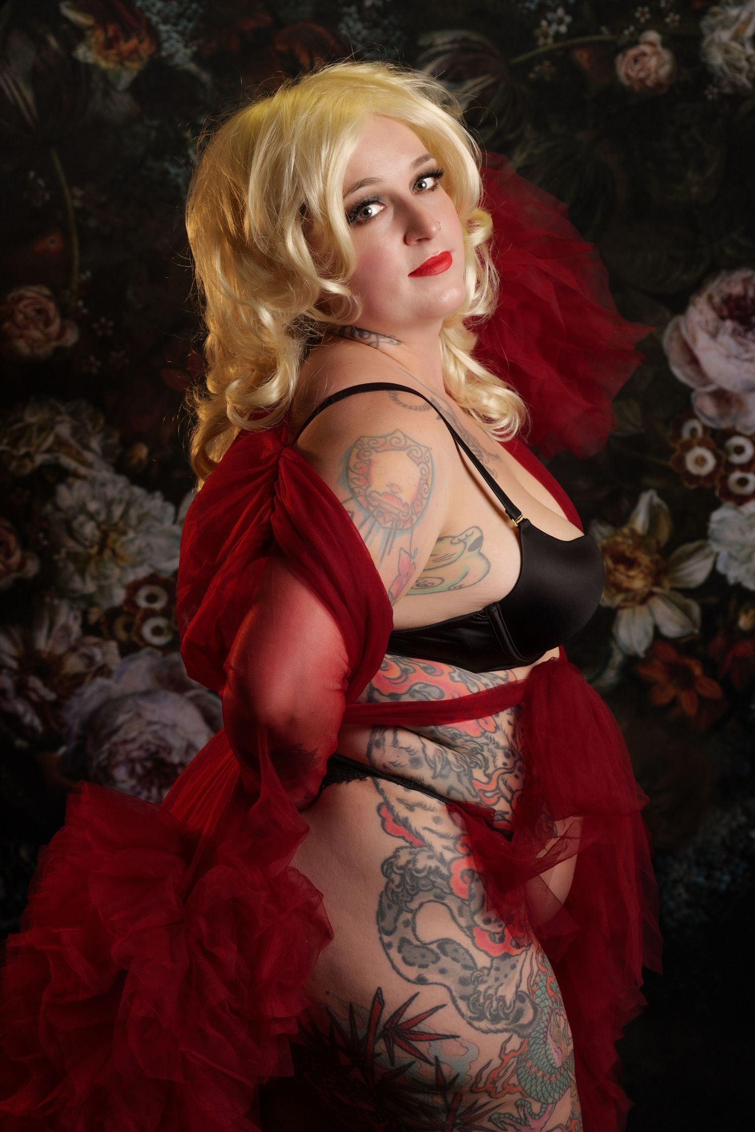 Tattooed blonde woman in black lingerie and red tulle robe posing against a dark floral backdrop.
