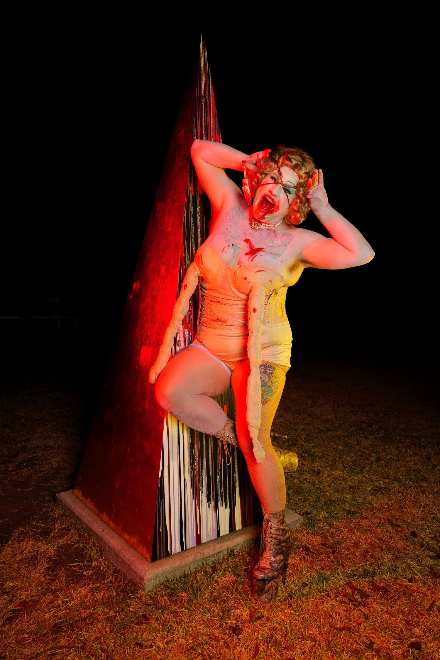 A woman covered in red paint screams dramatically against a red-lit triangular sculpture outdoors at night.