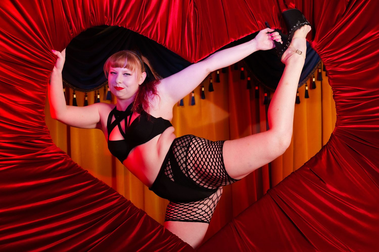 Flexible performer in black fishnet outfit poses dramatically against red heart-shaped curtain backdrop.