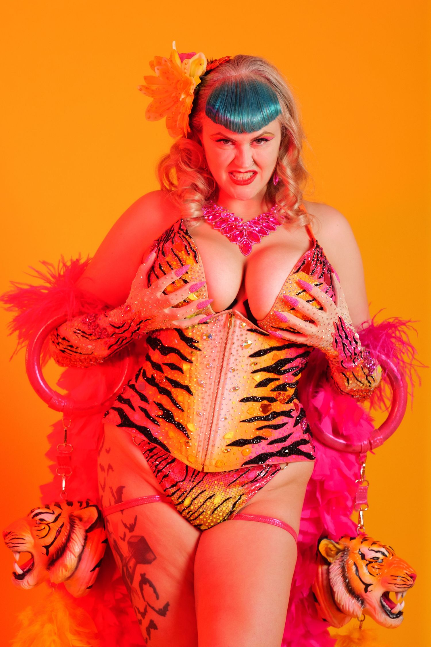 Woman in tiger-print corset and pink feather boa poses against bright orange background in colorful burlesque costume.