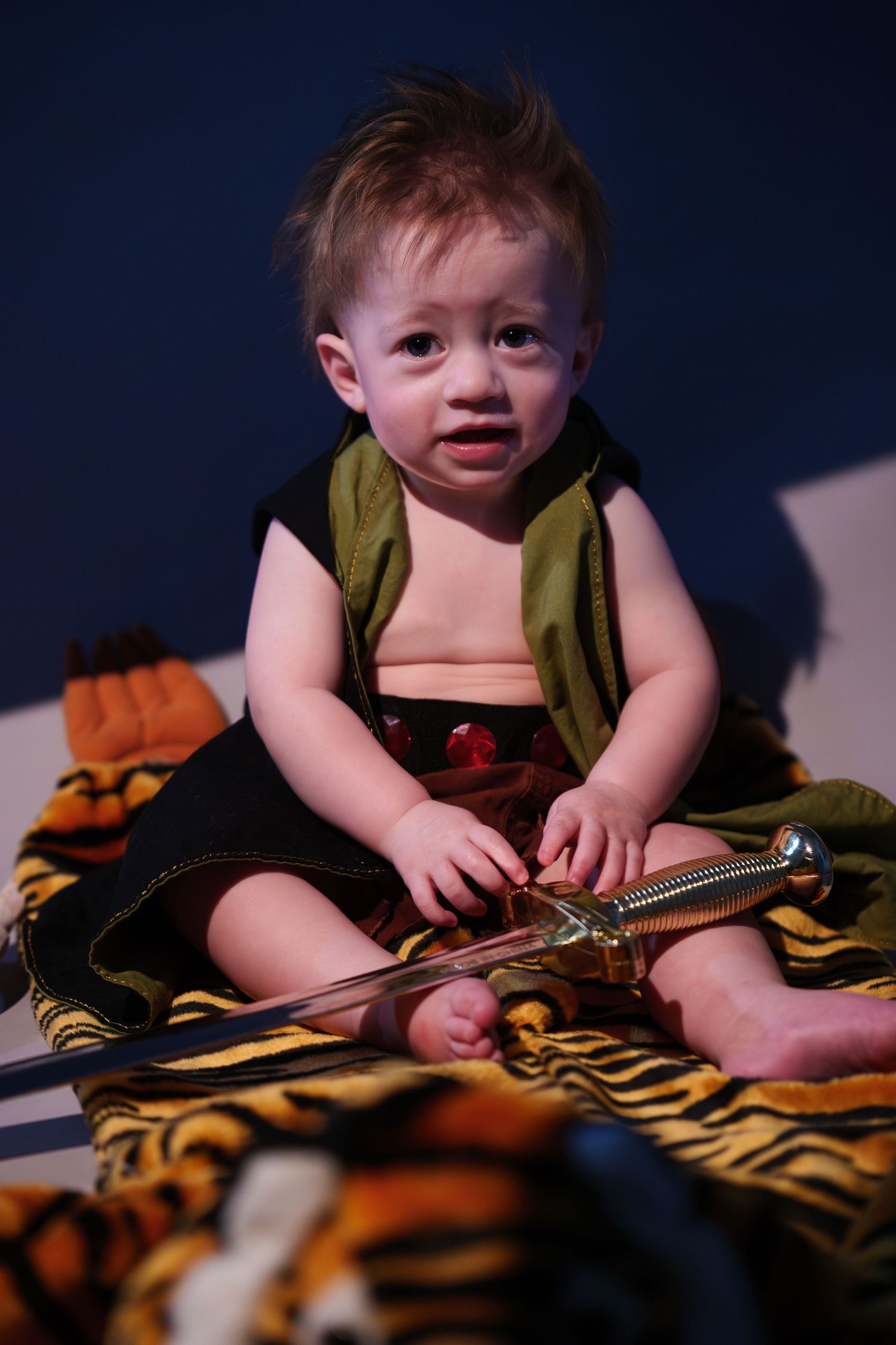 Baby dressed as a warrior sitting on tiger print blanket holding a toy sword in a dramatic studio setting.