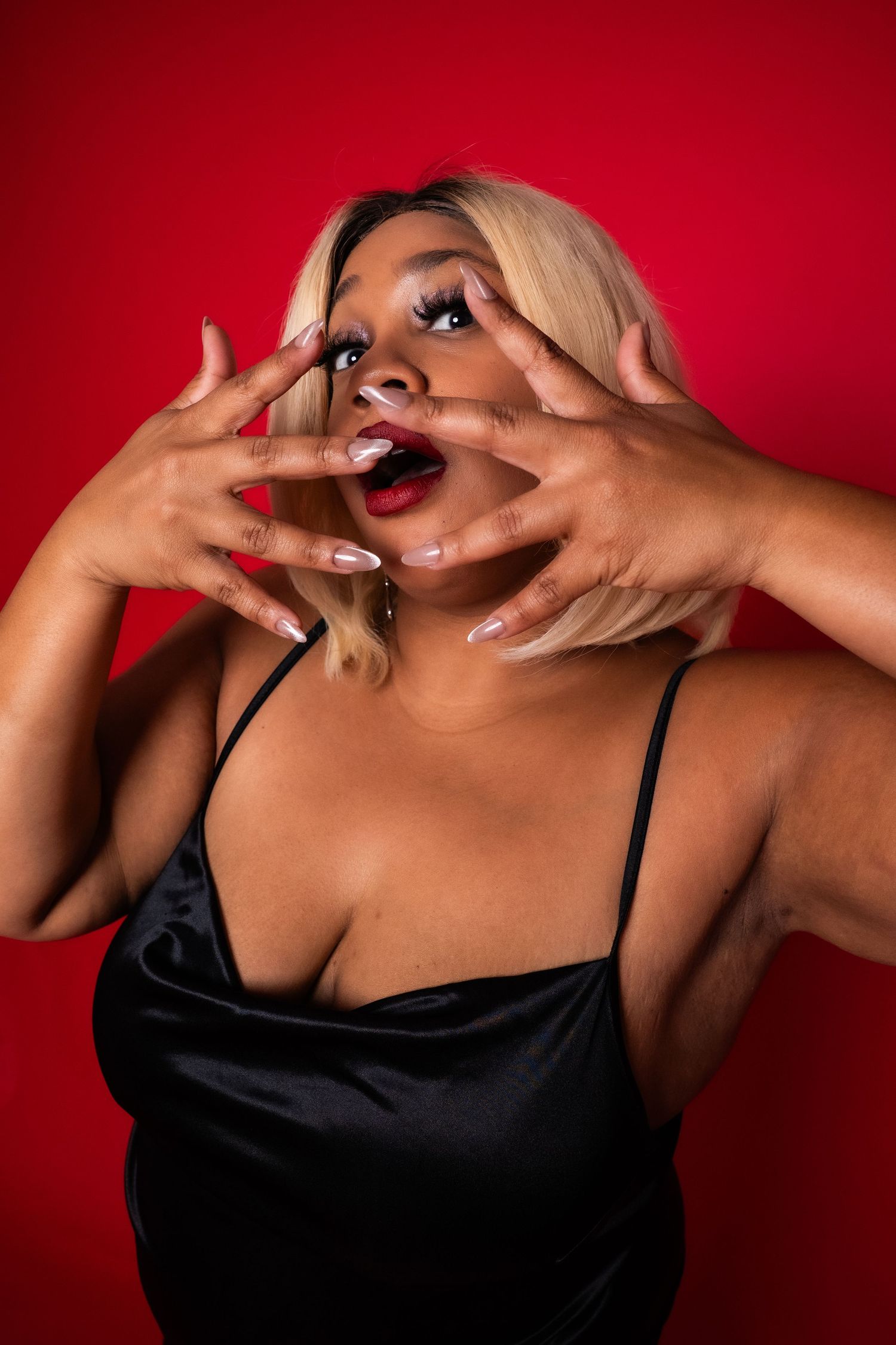 Expressive Black woman with blonde bob and red lips framing face with hands on red background.