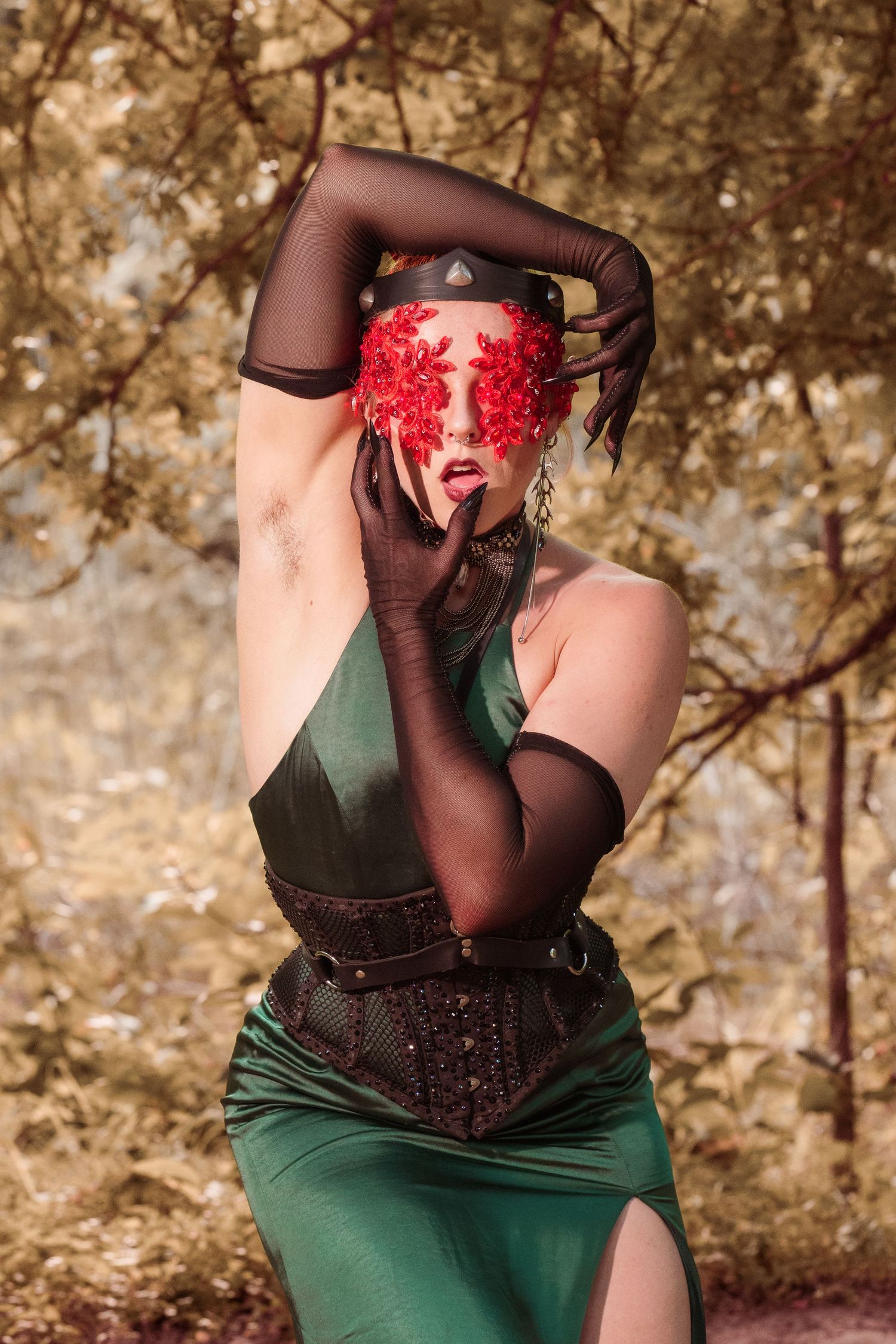 Woman in green dress and corset with red floral eye mask poses dramatically in golden forest wearing black mesh gloves.