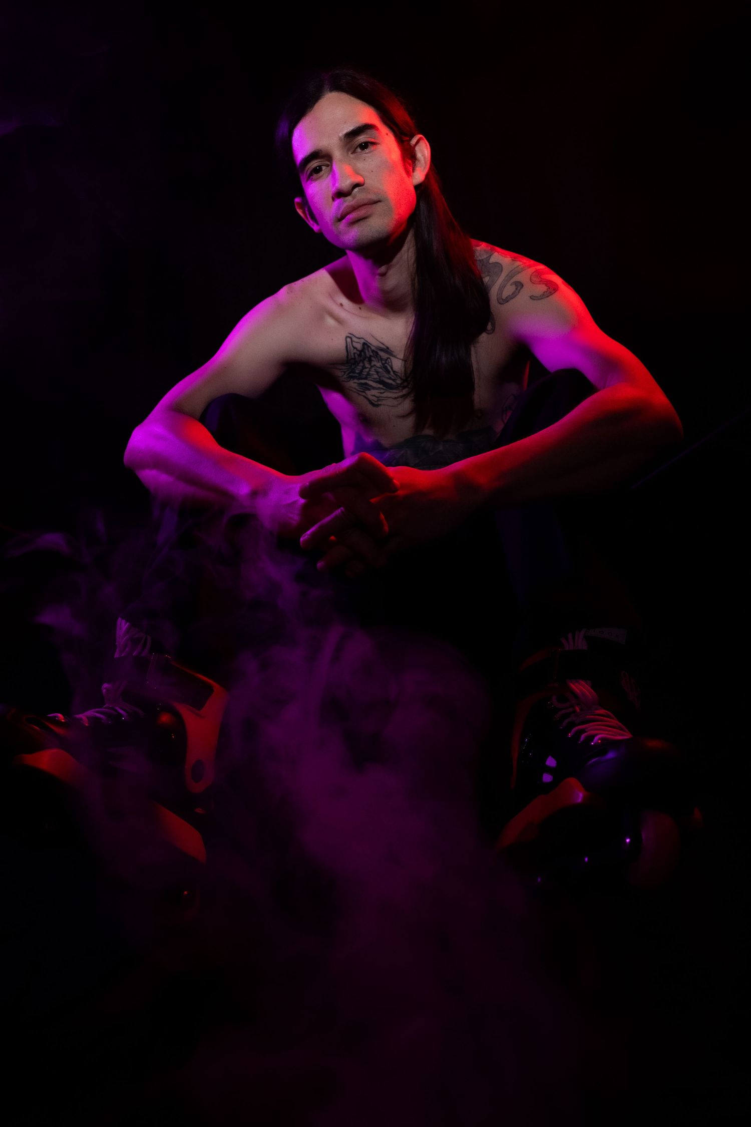 A shirtless tattooed man with long dark hair poses in moody purple and red neon lighting with smoke.