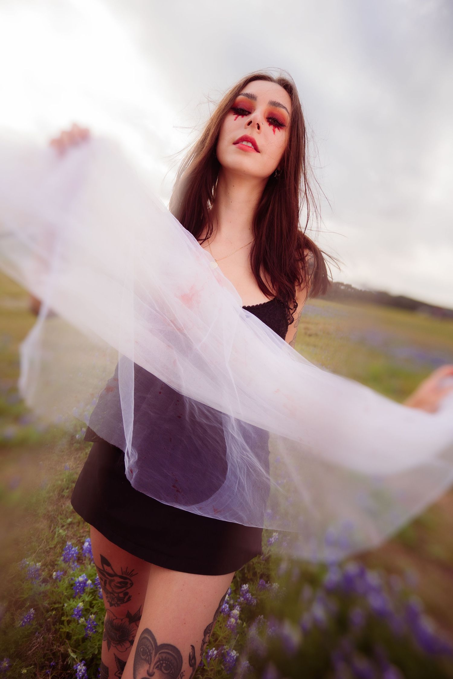 Tattooed woman with dramatic red eye makeup holds sheer white fabric in a flower field.