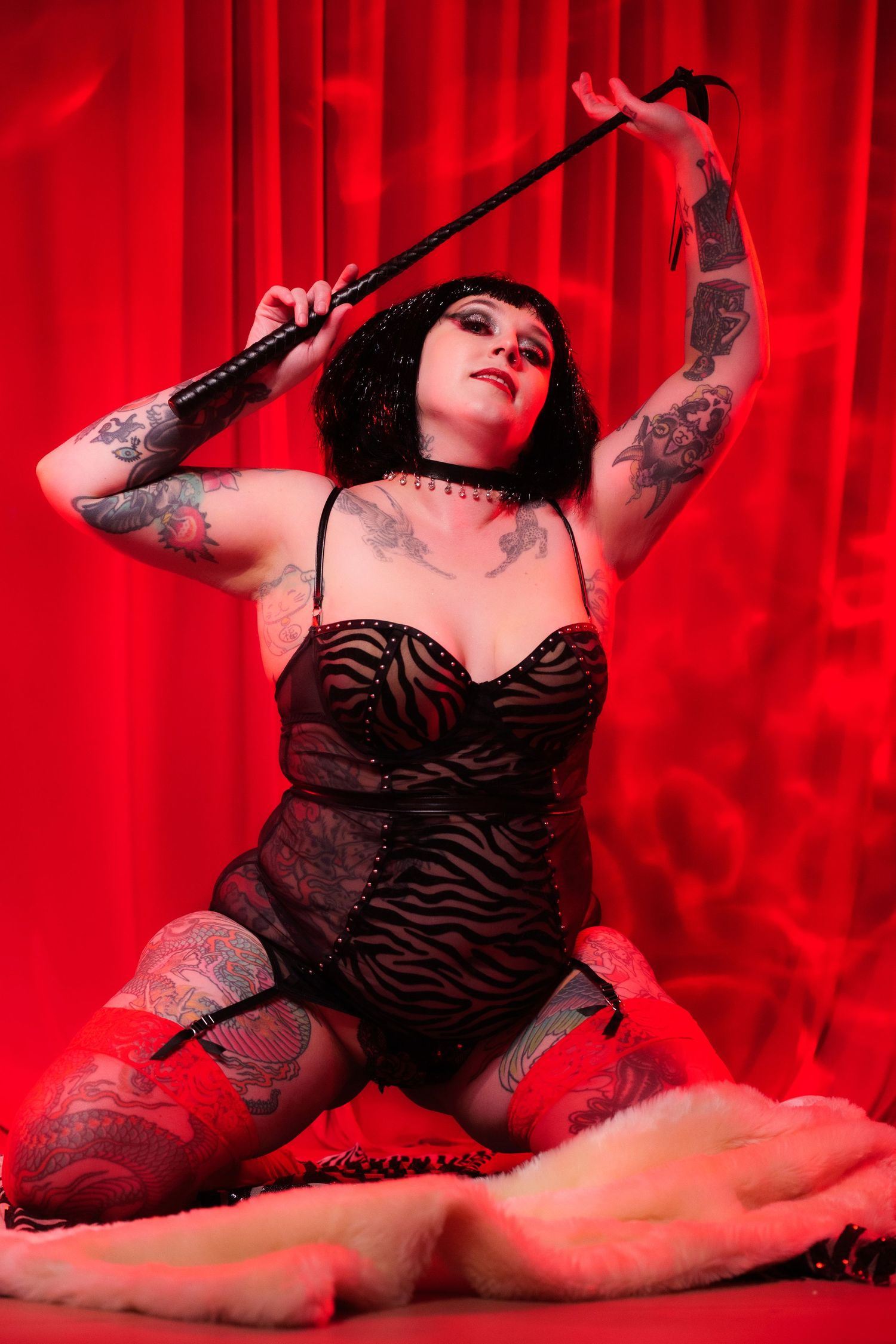 Tattooed woman in black lingerie and stockings posing with a riding crop against a red curtain backdrop.