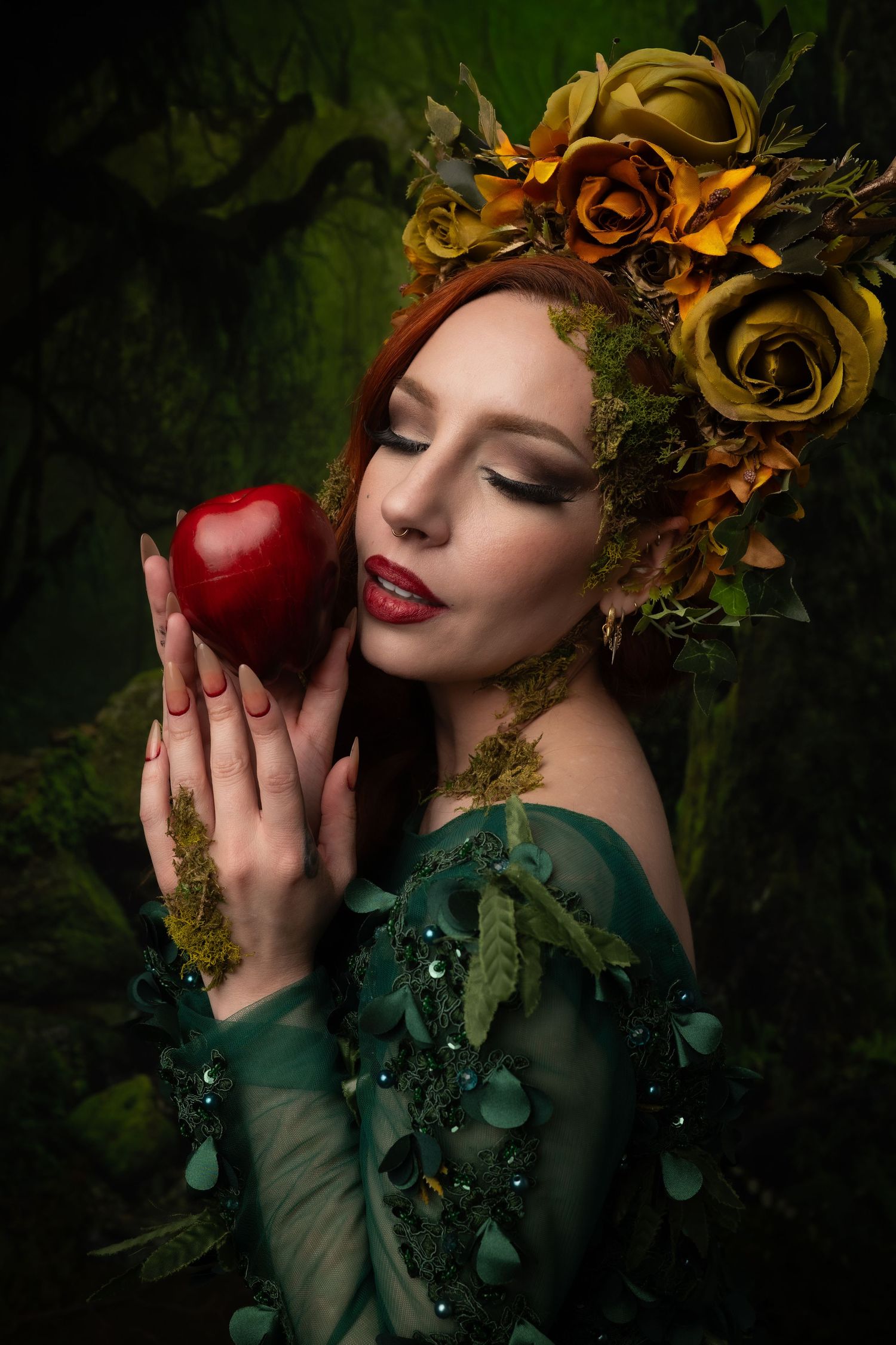A woman in a green floral gown holds a red apple, wearing a yellow rose crown in a dark enchanted forest setting.