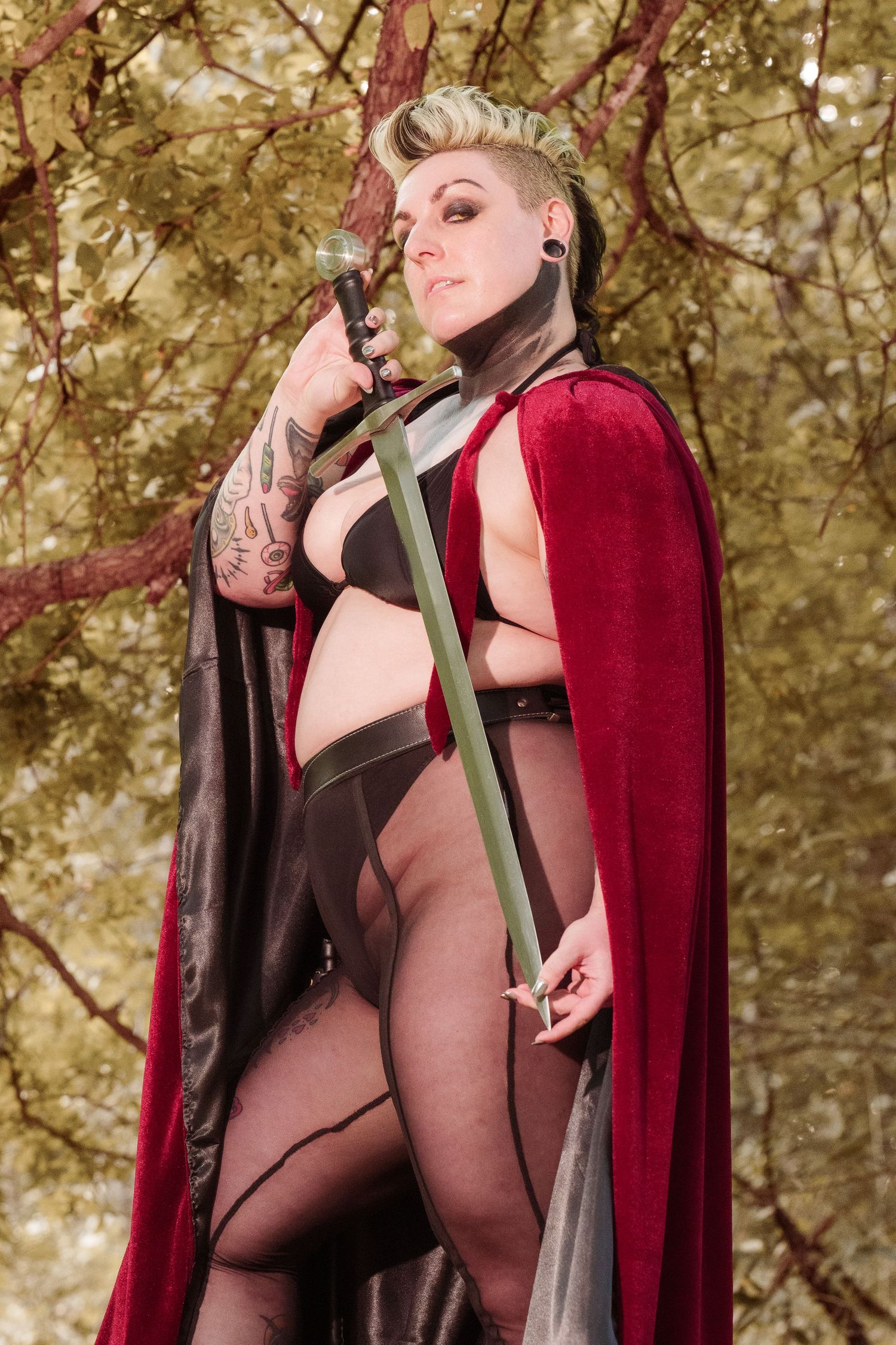 Tattooed cosplayer in red cape holds sword outdoors among green trees in fantasy warrior costume.