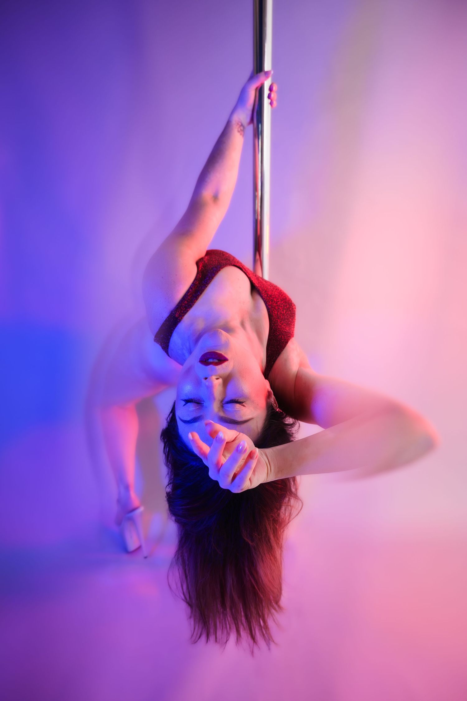Woman performing an inverted pole dance move under colorful purple and pink studio lighting.