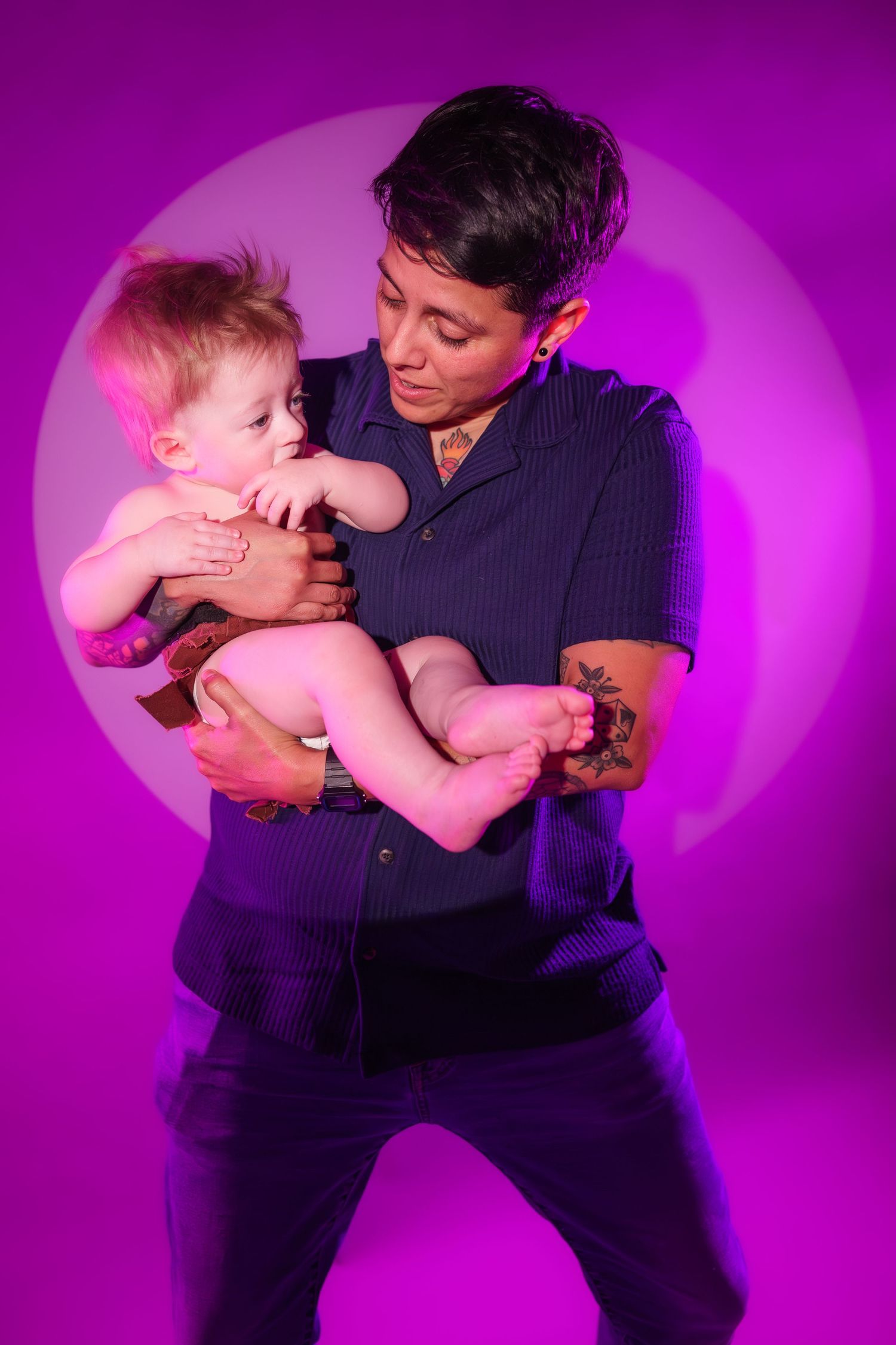 Person with short hair and tattoos holds a baby against a vibrant purple studio background.