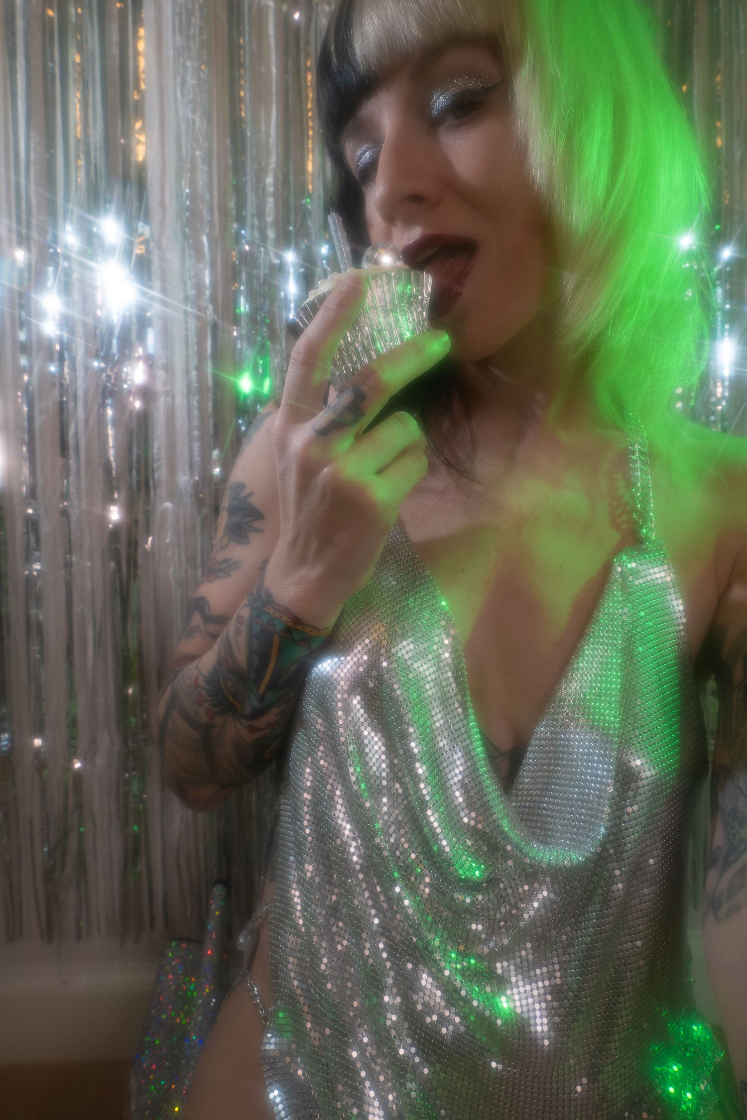Tattooed performer in silver sequin dress and green wig singing into microphone under sparkling green stage lights.