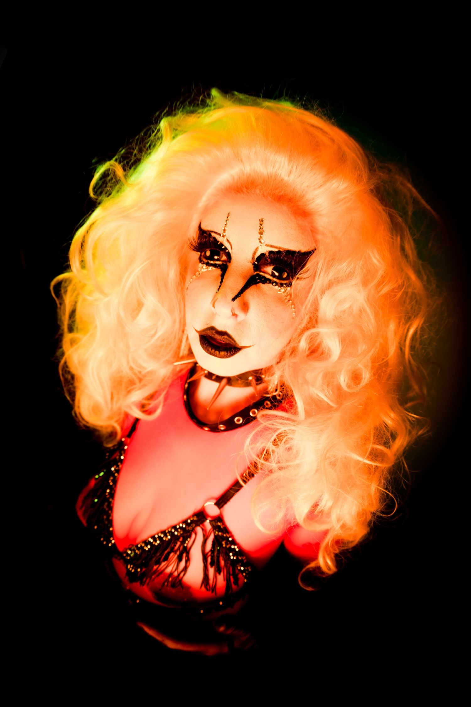 Drag performer with voluminous blonde wig and dramatic gothic makeup illuminated by warm orange light on black background.