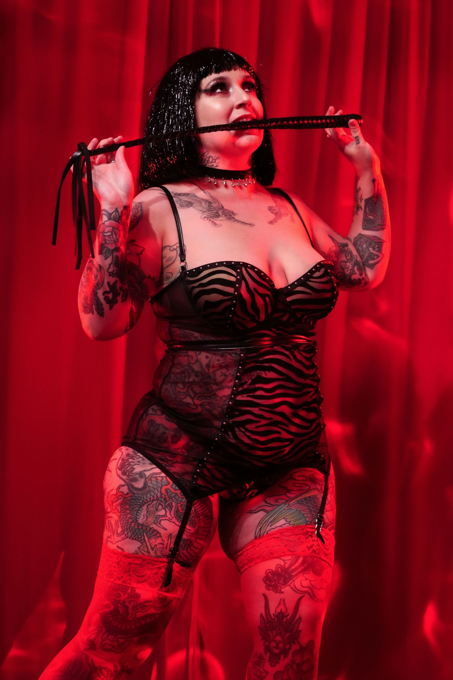 Tattooed woman in red and black lingerie with stockings poses against red curtain backdrop.