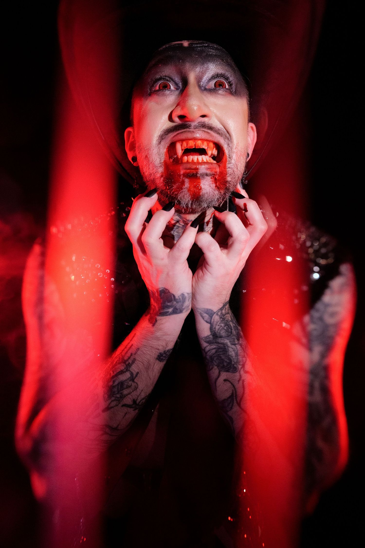 Tattooed man with vampire fangs and fake blood on face, illuminated by dramatic red light in dark setting.