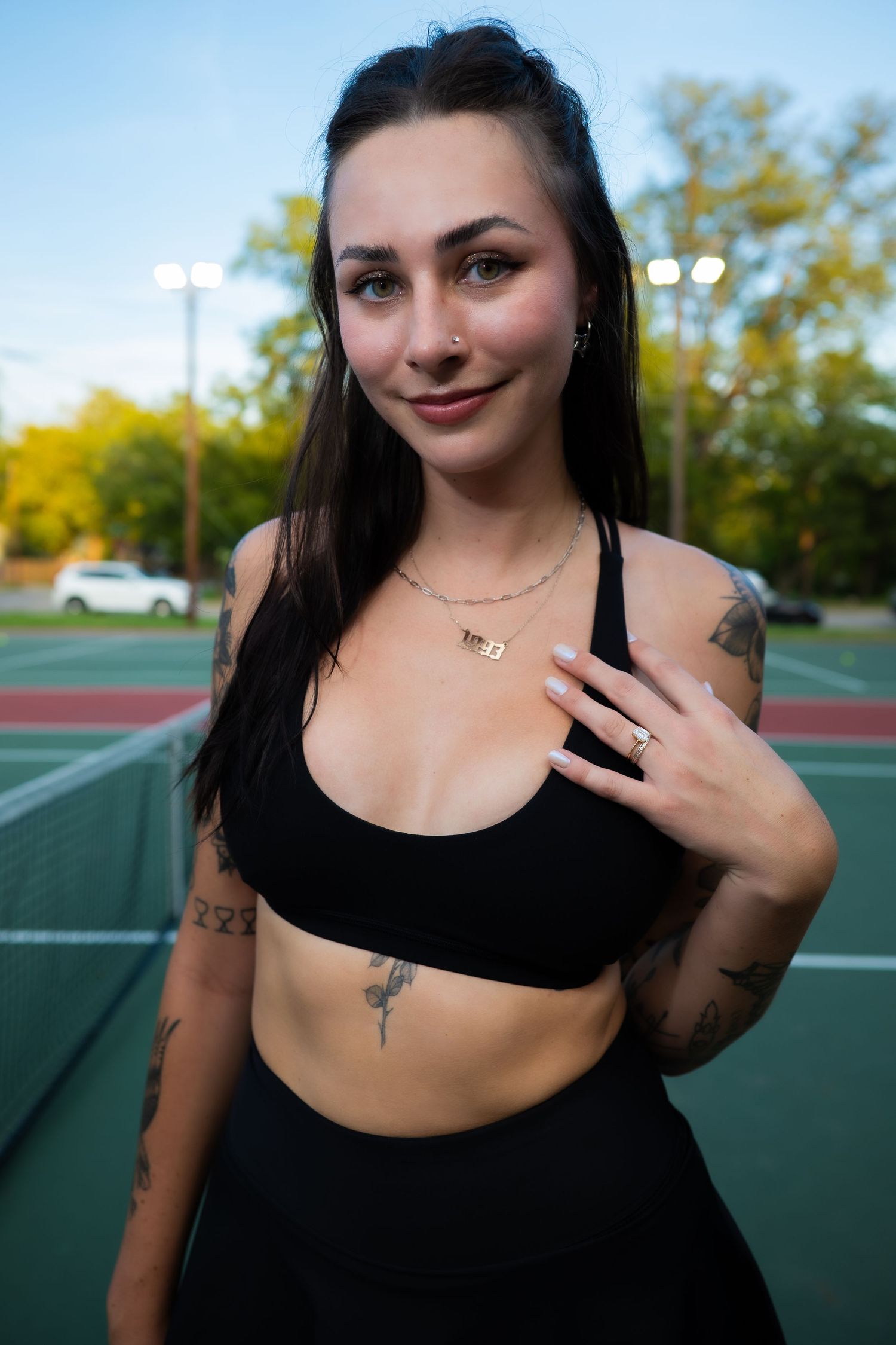 Smiling tattooed brunette woman in black sports bra on outdoor tennis court.