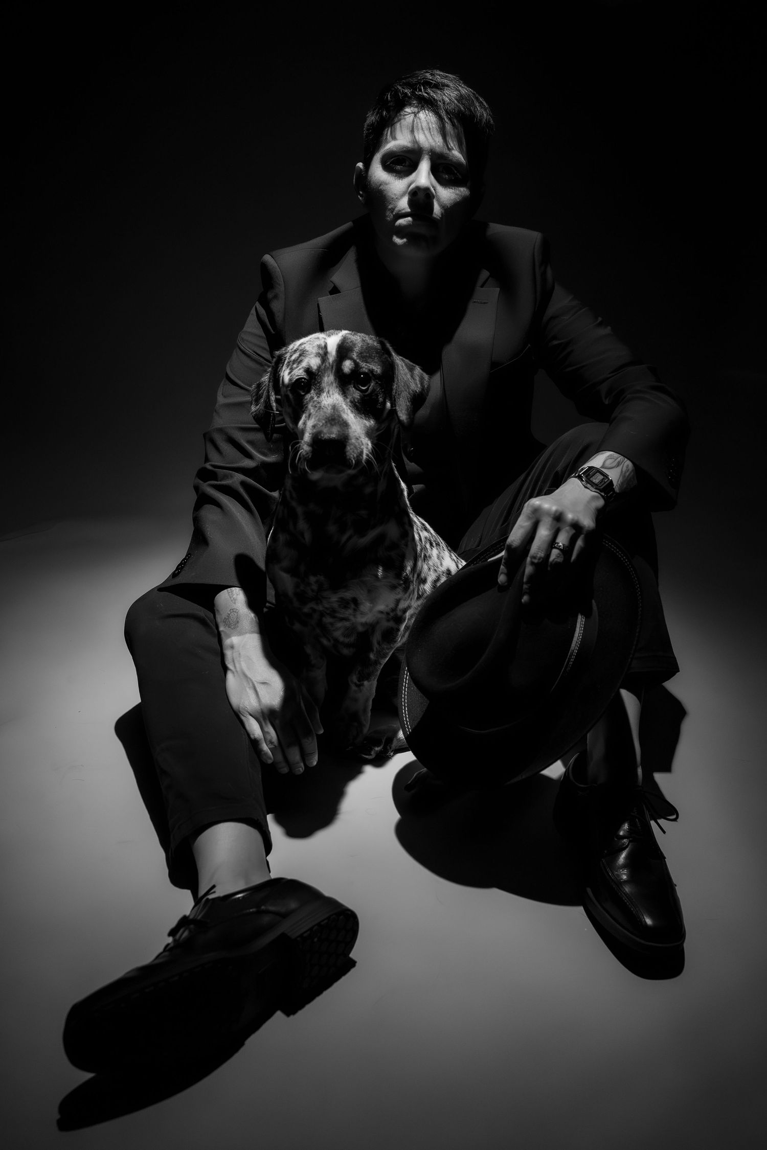 A well-dressed man in a black suit sits beside a Dalmatian dog in a dramatic black and white portrait.