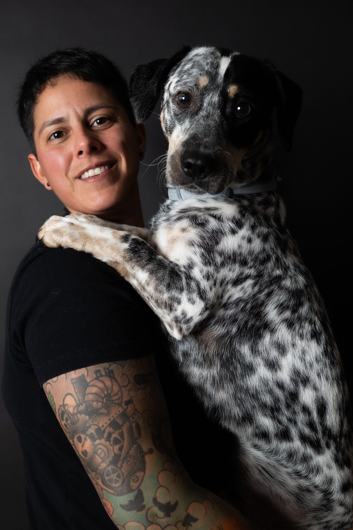 Smiling woman with tattoos and short hair holds a spotted Dalmatian dog against dark background.