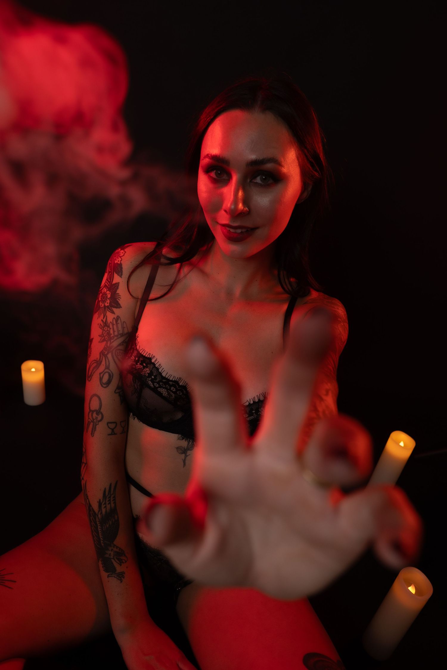 Tattooed woman in black lace lingerie reaches toward camera surrounded by candles in moody red-lit dark setting.