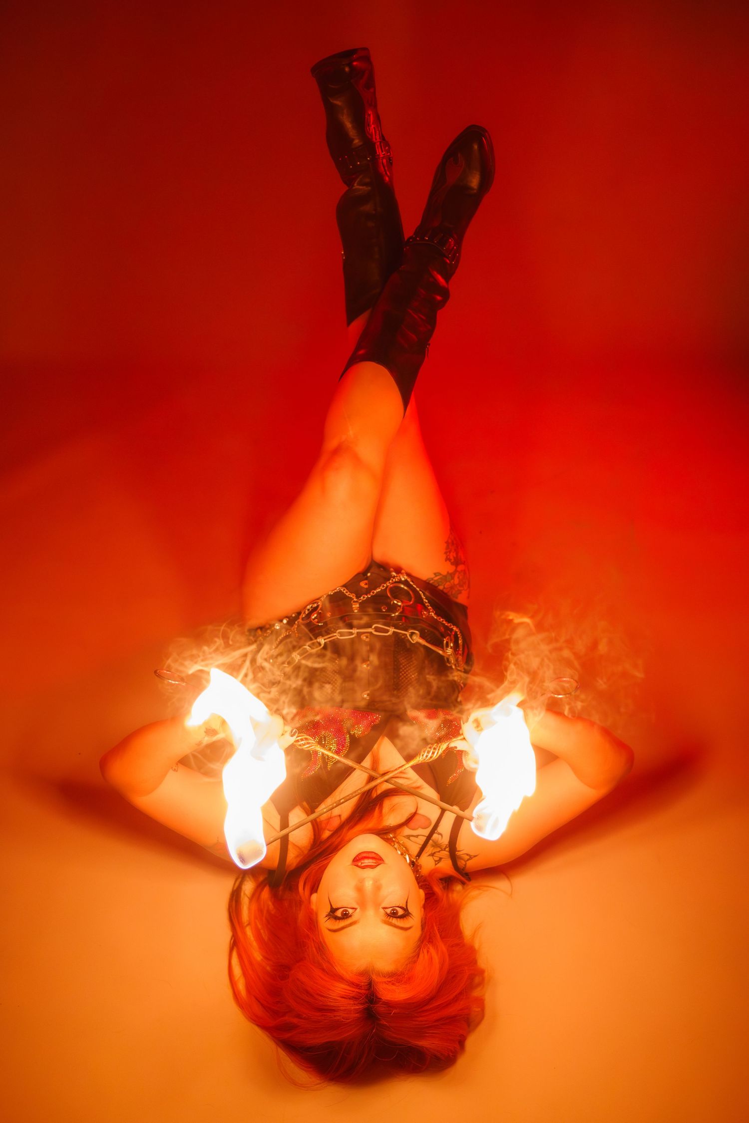 Red-haired woman holds fire poi while lying on her back in a fiery red-lit studio.