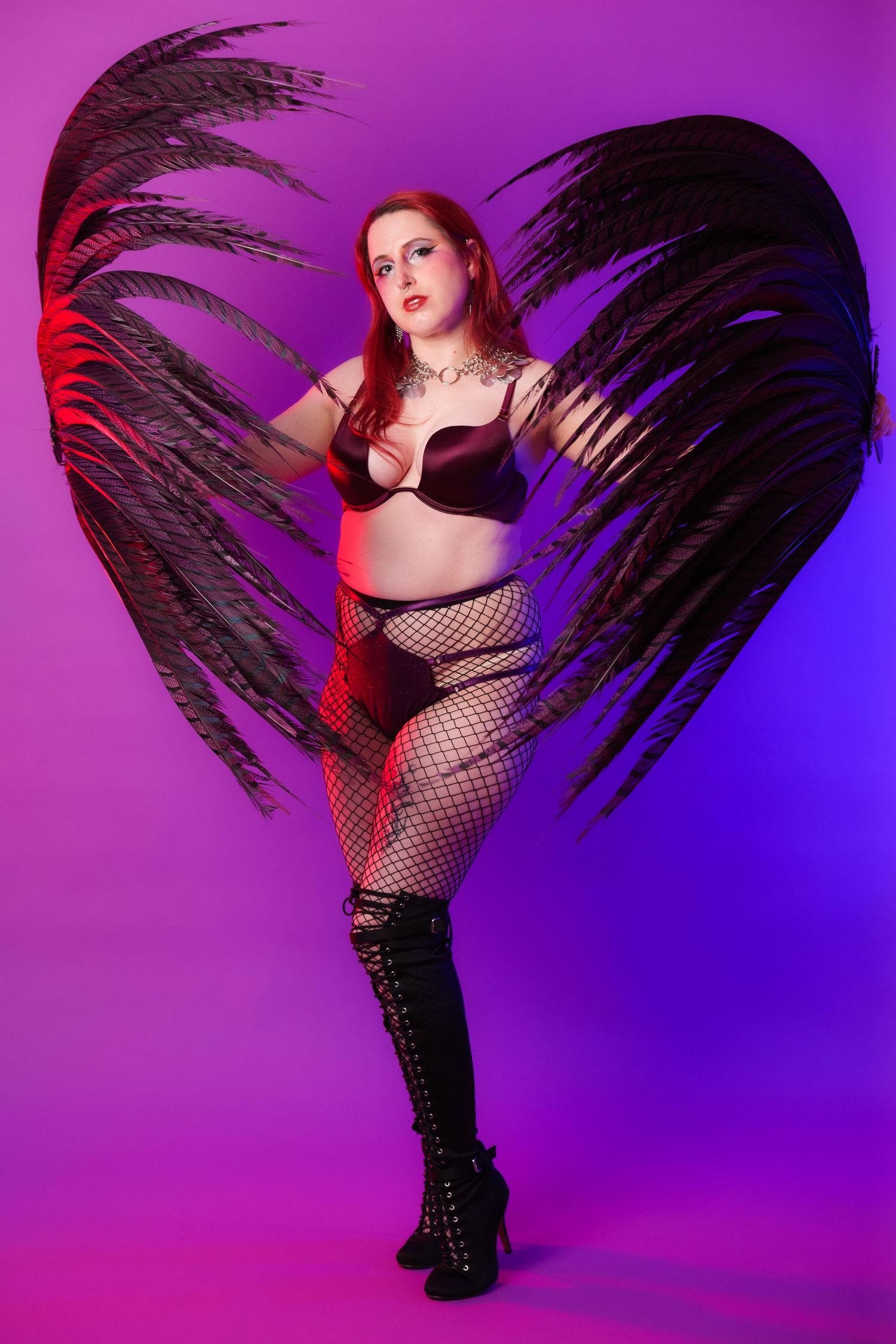 Red-haired woman in black lingerie and fishnet stockings poses with large dark feathered wings on purple background.