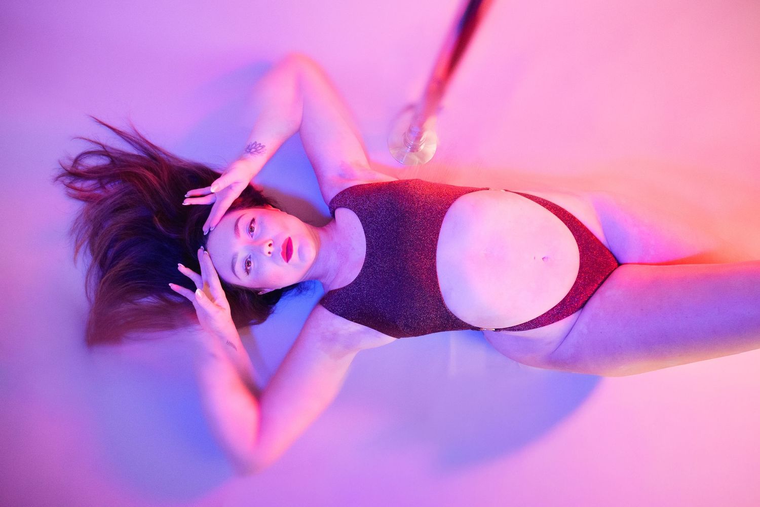 Woman in dark swimsuit poses under vibrant pink and purple neon lighting in artistic fashion photography.