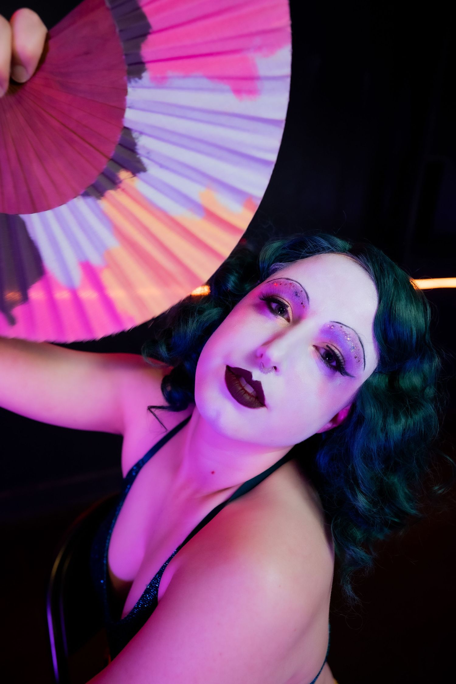 Performer with purple makeup holding colorful fan under pink neon light.
