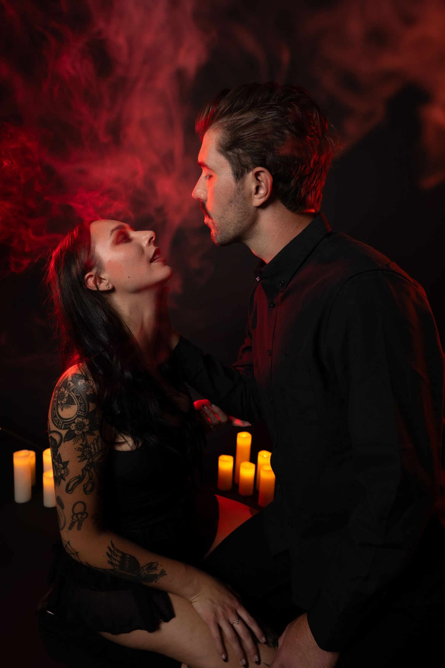 Man and woman in gothic costumes pose dramatically with candles and red smoke in dark setting.