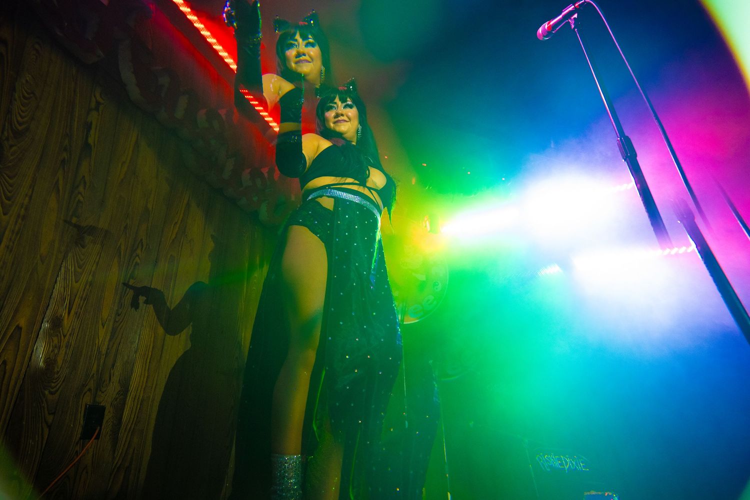 Two performers in cat-eye makeup and revealing outfits pose on stage under vibrant colorful concert lighting.