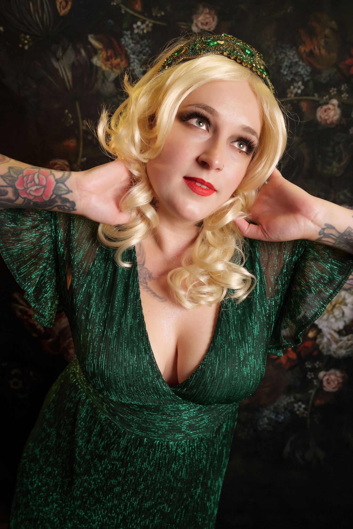 Blonde woman in green glittery deep-V dress with jeweled headband and tattoos poses against floral backdrop.