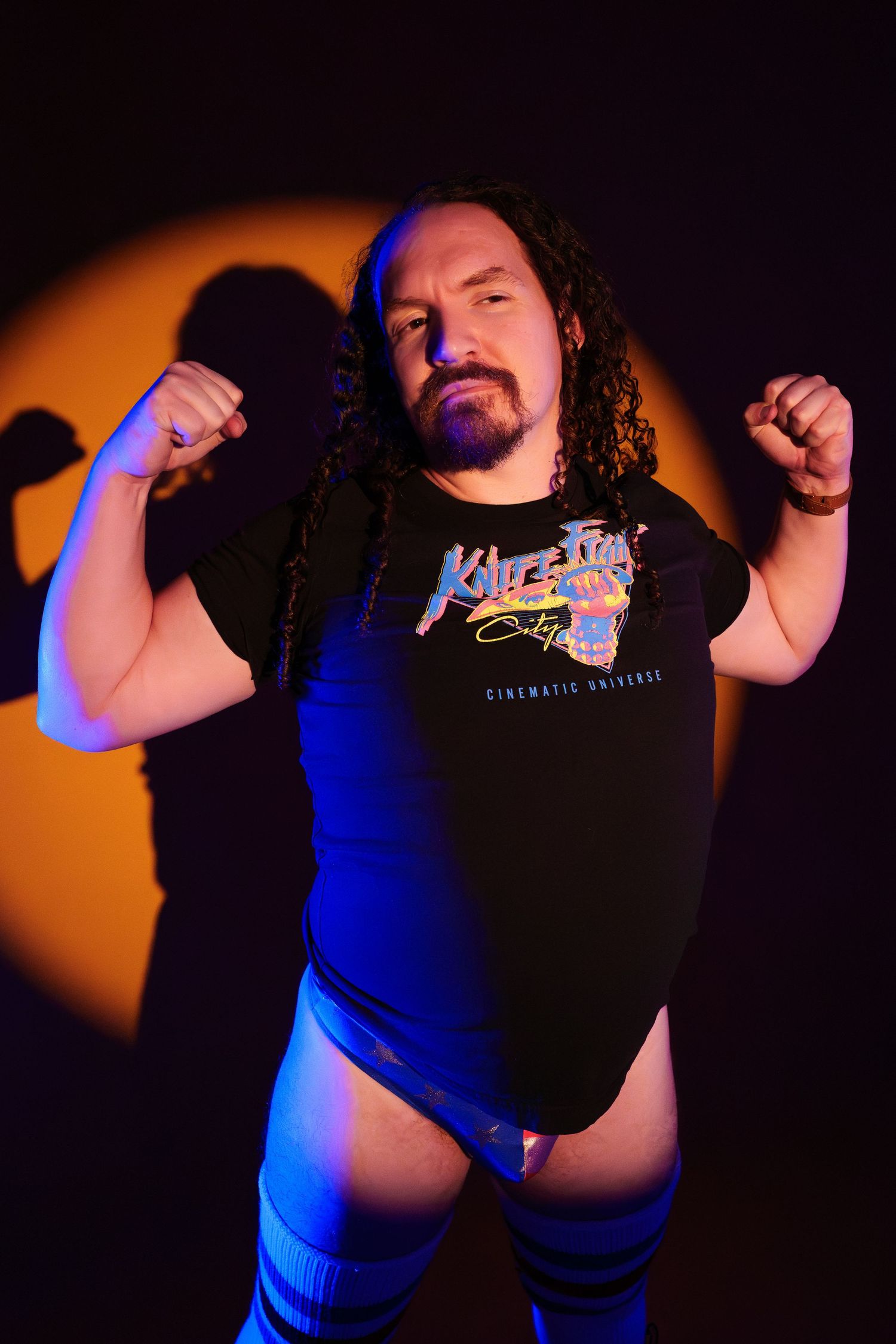 Muscular wrestler with long curly hair flexes arms wearing a black graphic tee under dramatic orange and blue stage lighting.