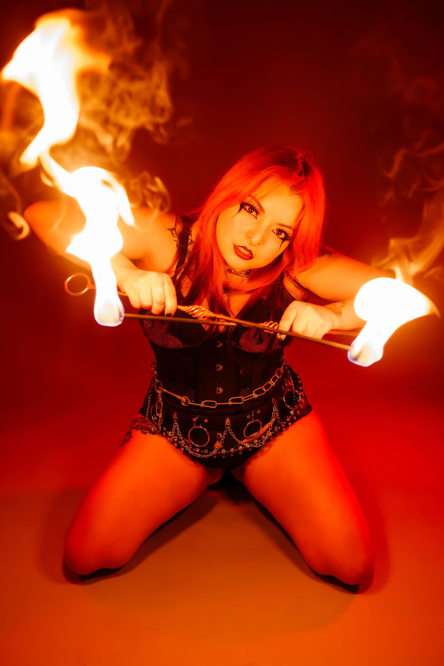 Red-haired fire performer kneels dramatically wielding flaming poi in a red-lit studio.