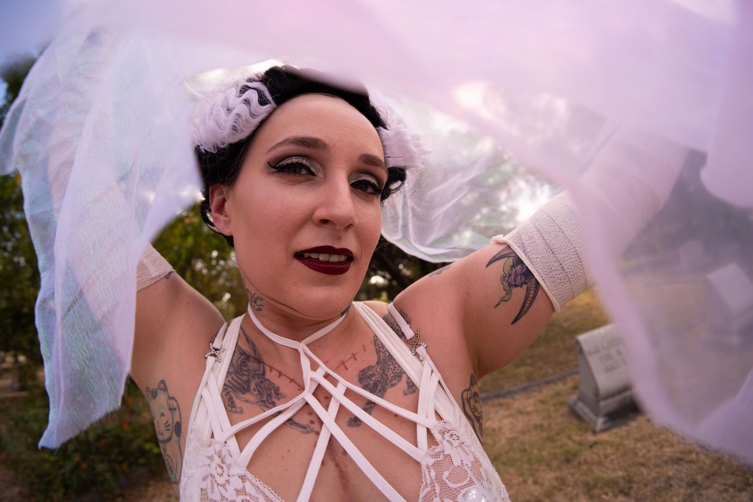 Tattooed bride in white lace dress with veil in cemetery.