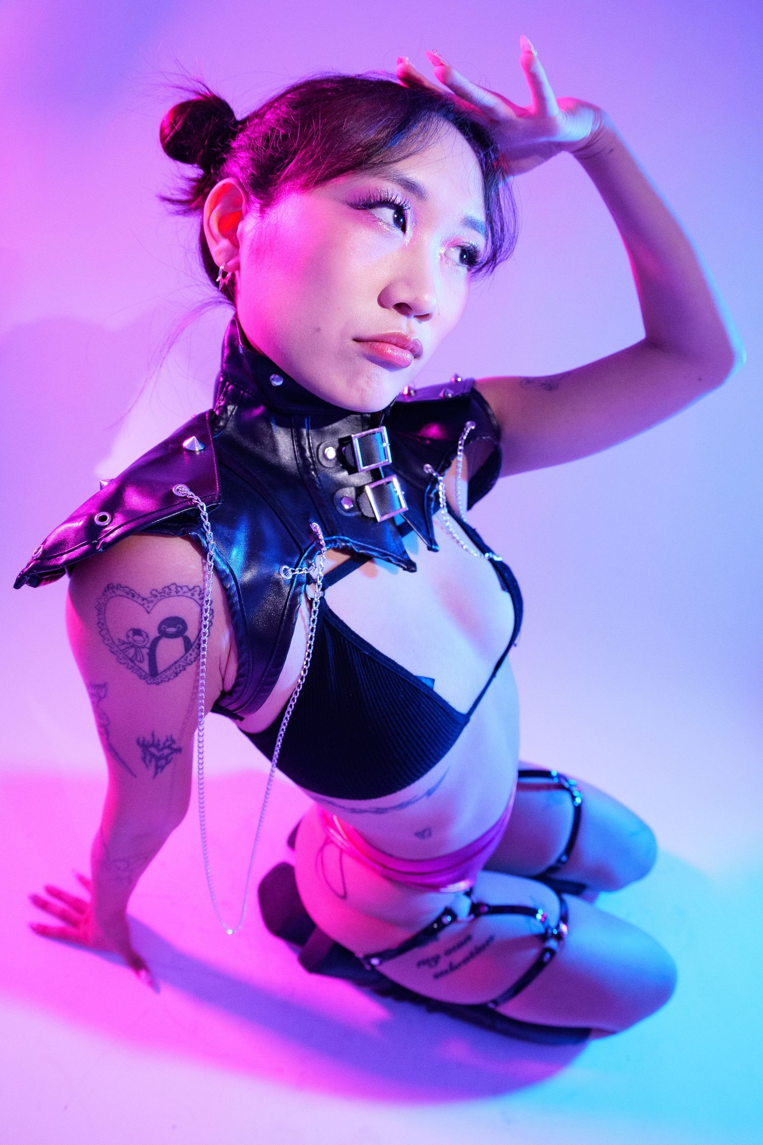 Young woman in edgy black leather harness outfit with tattoos posing under pink and purple neon studio lighting.