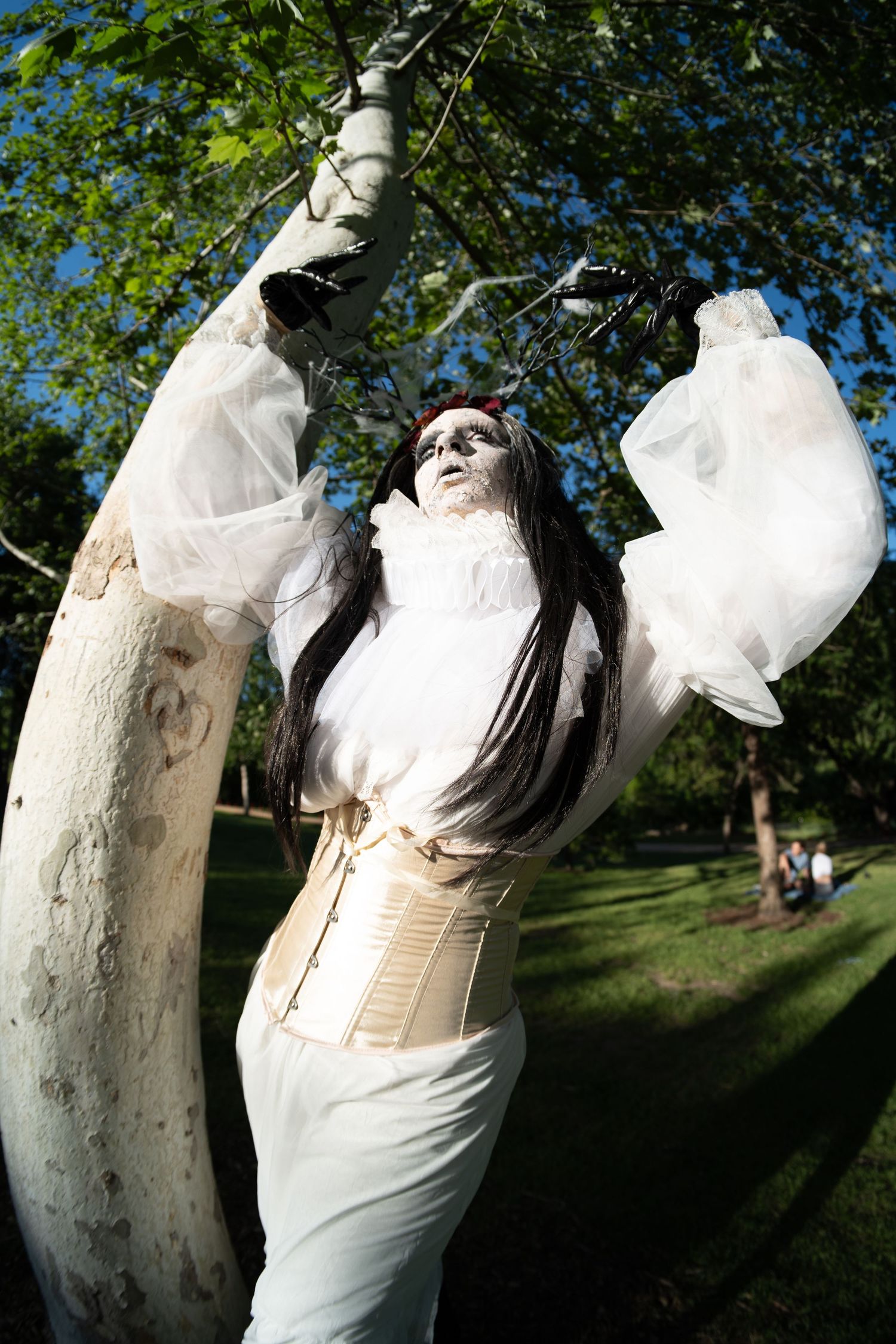 Woman in white costume with dramatic makeup and corset poses between birch trees in sunny park.