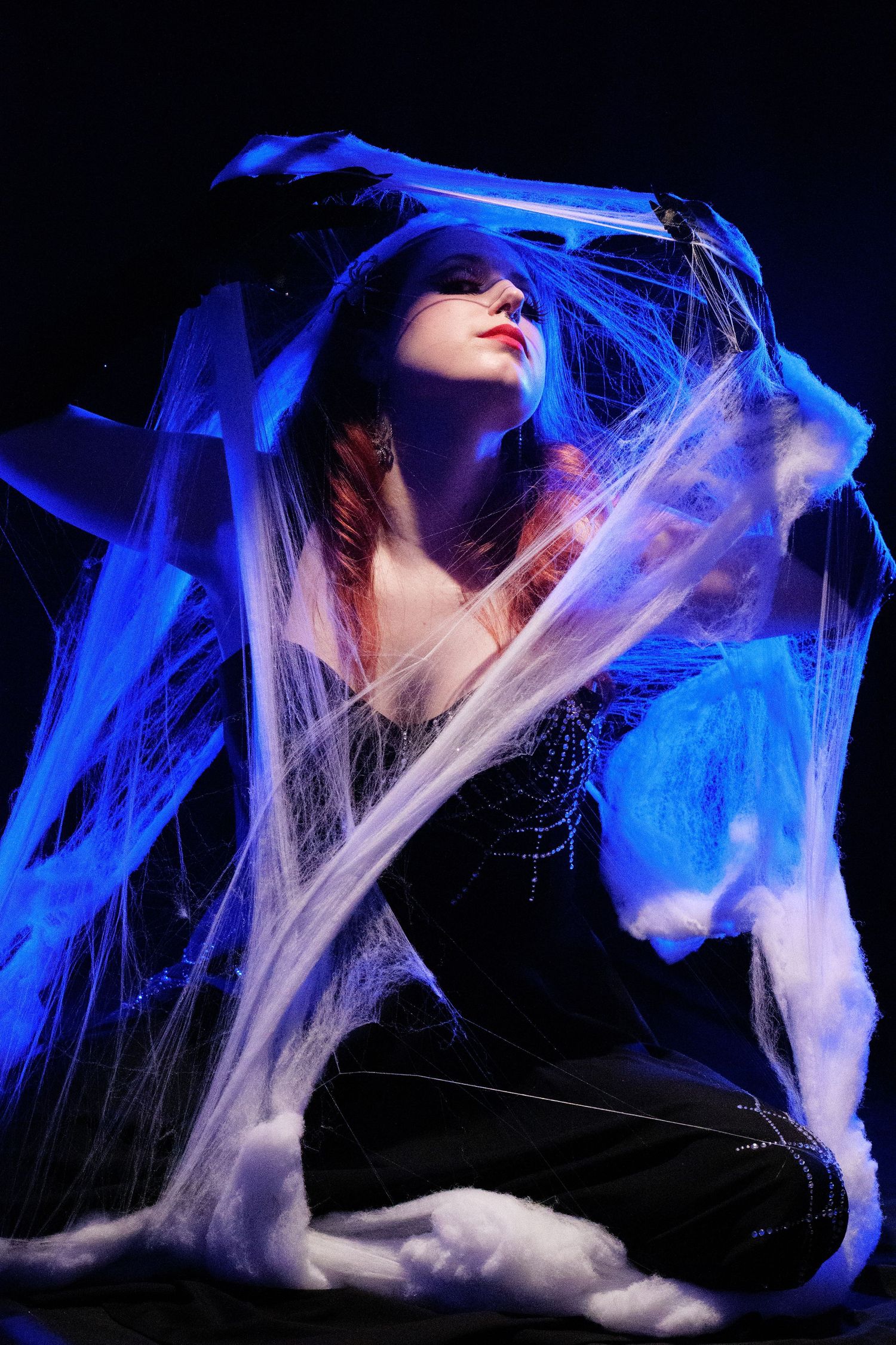 A woman draped in white spider webs poses under dramatic blue lighting against a dark background.